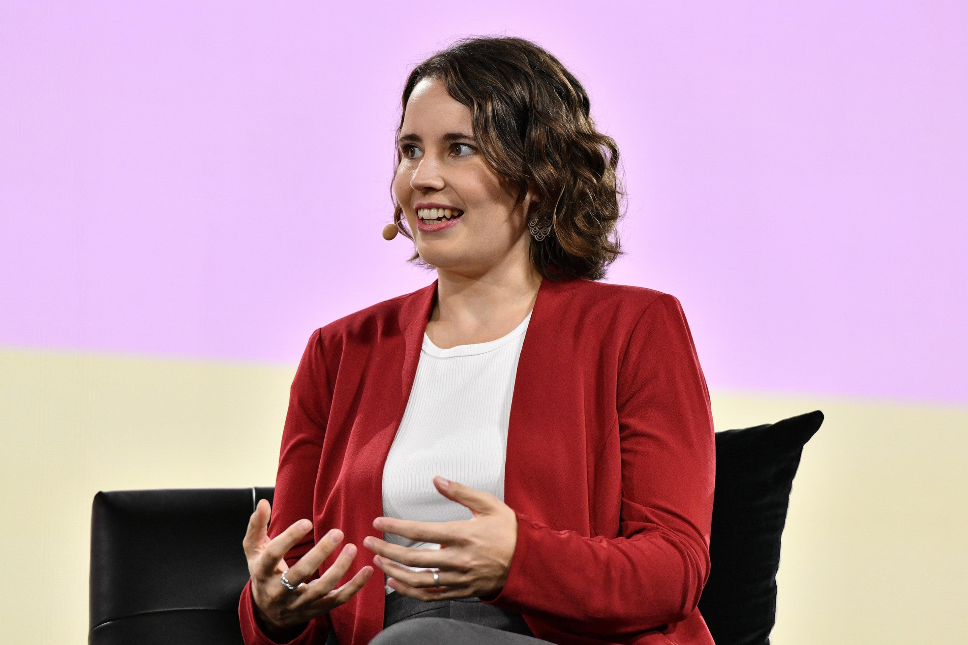 Helen Toner of Georgetown's CSET wears a red cardigan and a white blouse speaking at Vox Media's 2023 Code Conference. Behind her is a pink and yellow background.