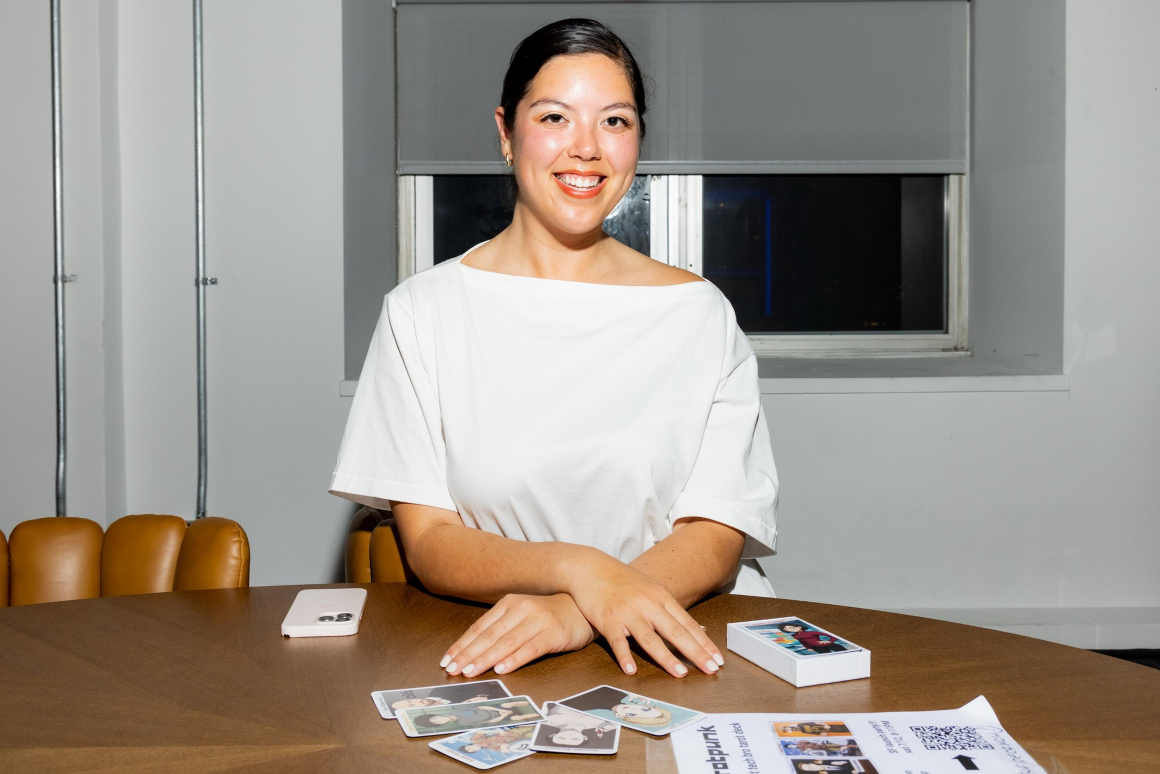 A person in a white shirt sits at a table with playing cards, a smartphone, and a small box. They are smiling, seated in a room with a window behind them.