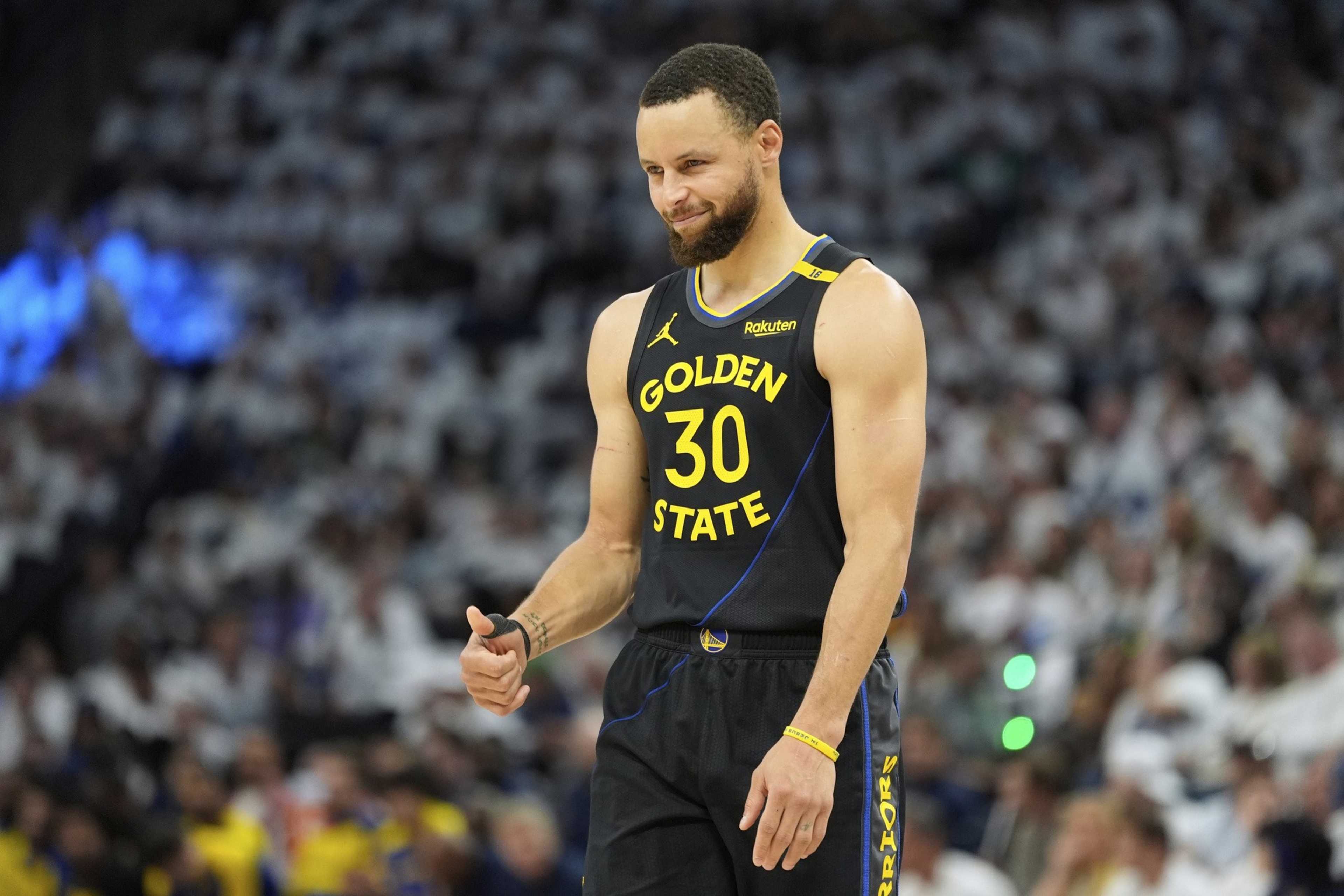 A basketball player in a black Golden State jersey (#30) gives a thumbs-up. He smiles slightly, standing on the court with a blurred crowd in the background.