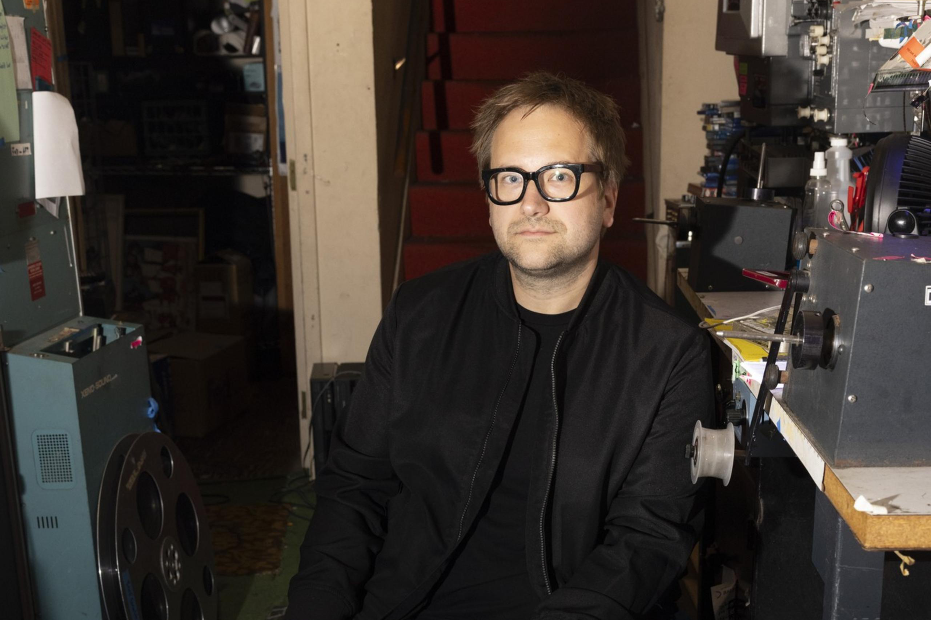 A man in a black shirt and jacket and gray pants sits facing the camera in a projection room surrounded by film reels and other materials.