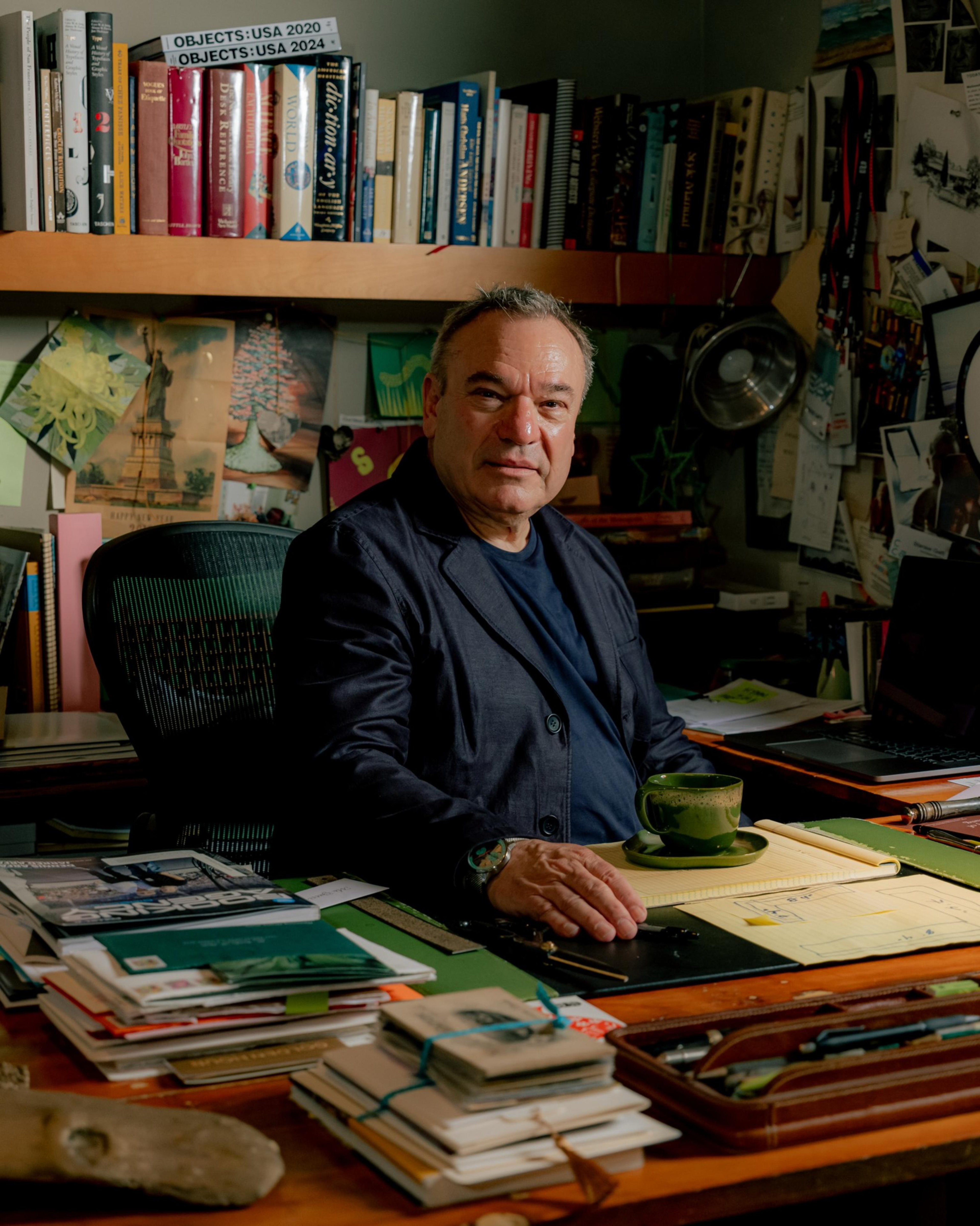 A man sits at a cluttered desk with books, papers, and a green cup while behind him shelves hold more books and pinned notes on walls.