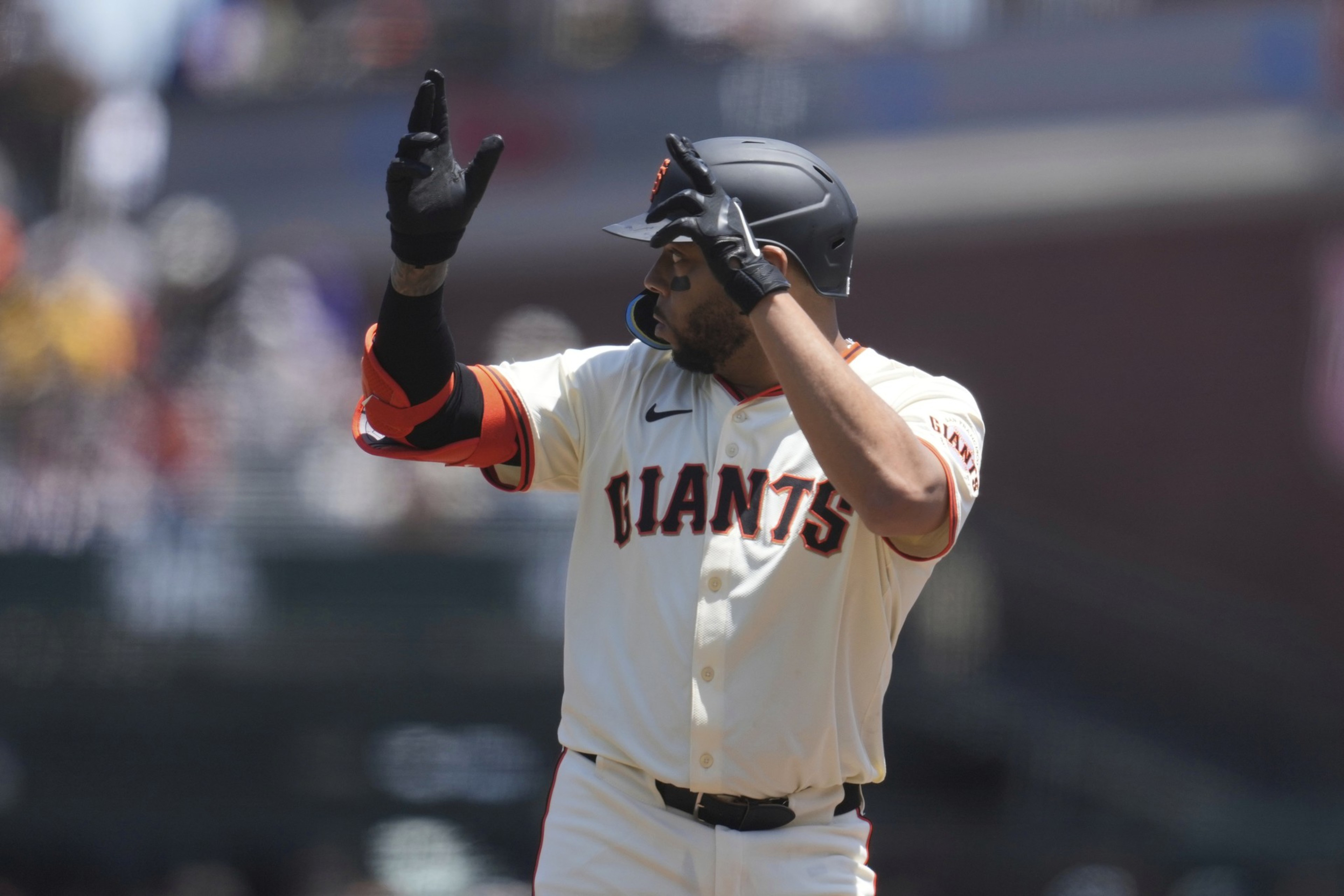 A baseball player wearing a Giants uniform and helmet stands on the field, gesturing with both gloved hands. The background is a blurred stadium.
