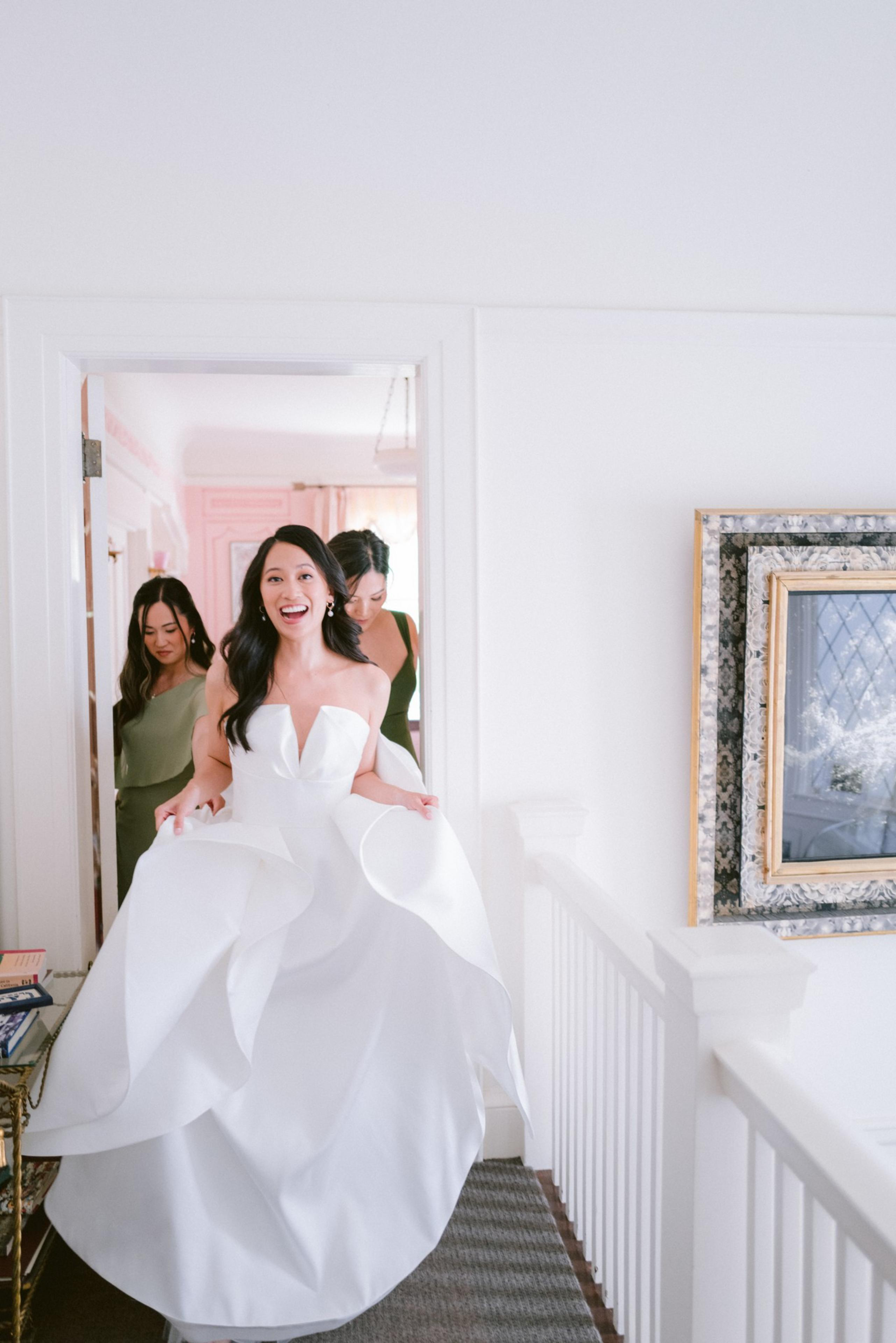 A joyful bride in a white strapless gown holds her dress while walking down a hallway, followed by two bridesmaids in olive dresses.