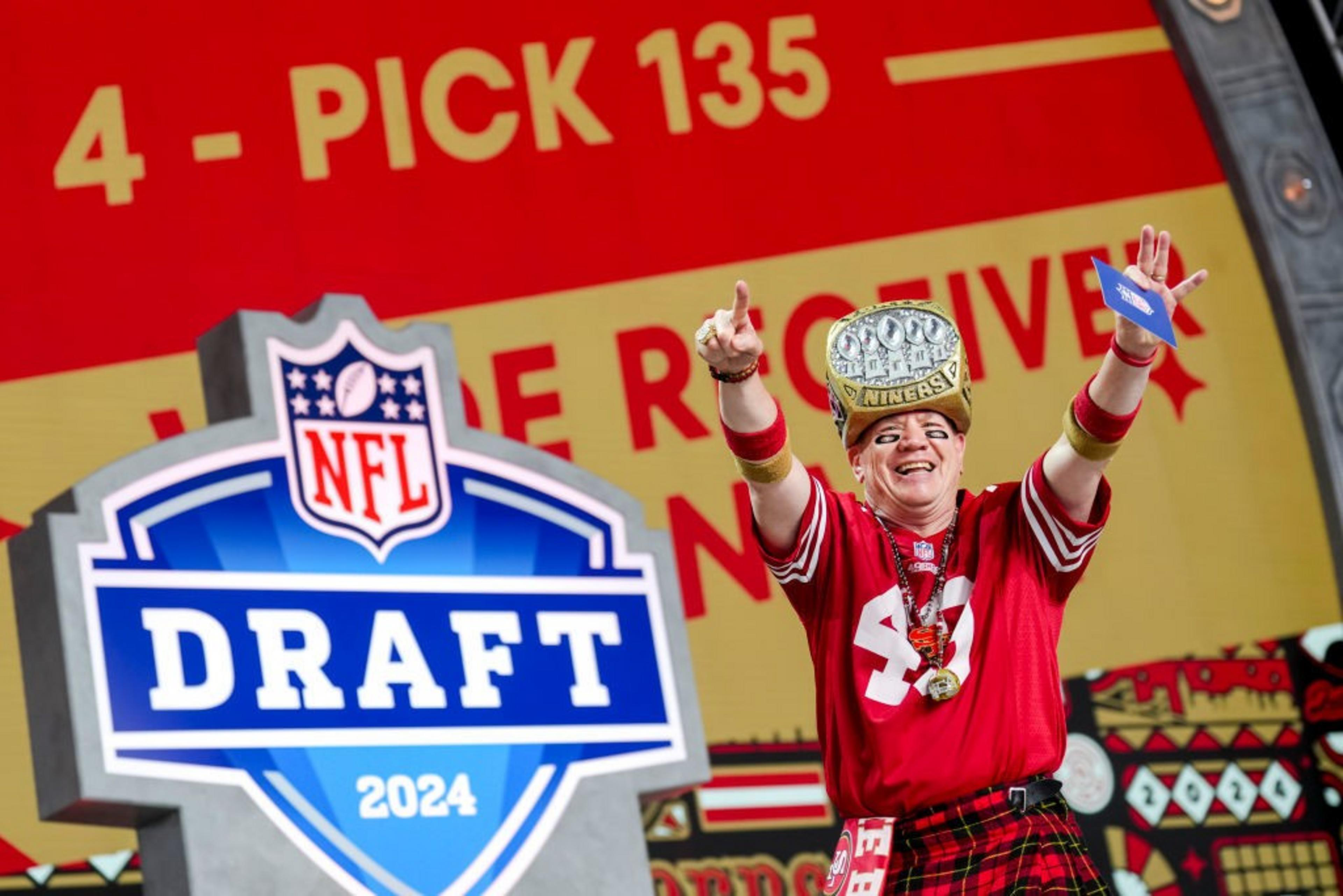 A smiling person in a red sports jersey and plaid pants cheers on stage at the NFL Draft 2024, wearing a large ring hat and holding a card.