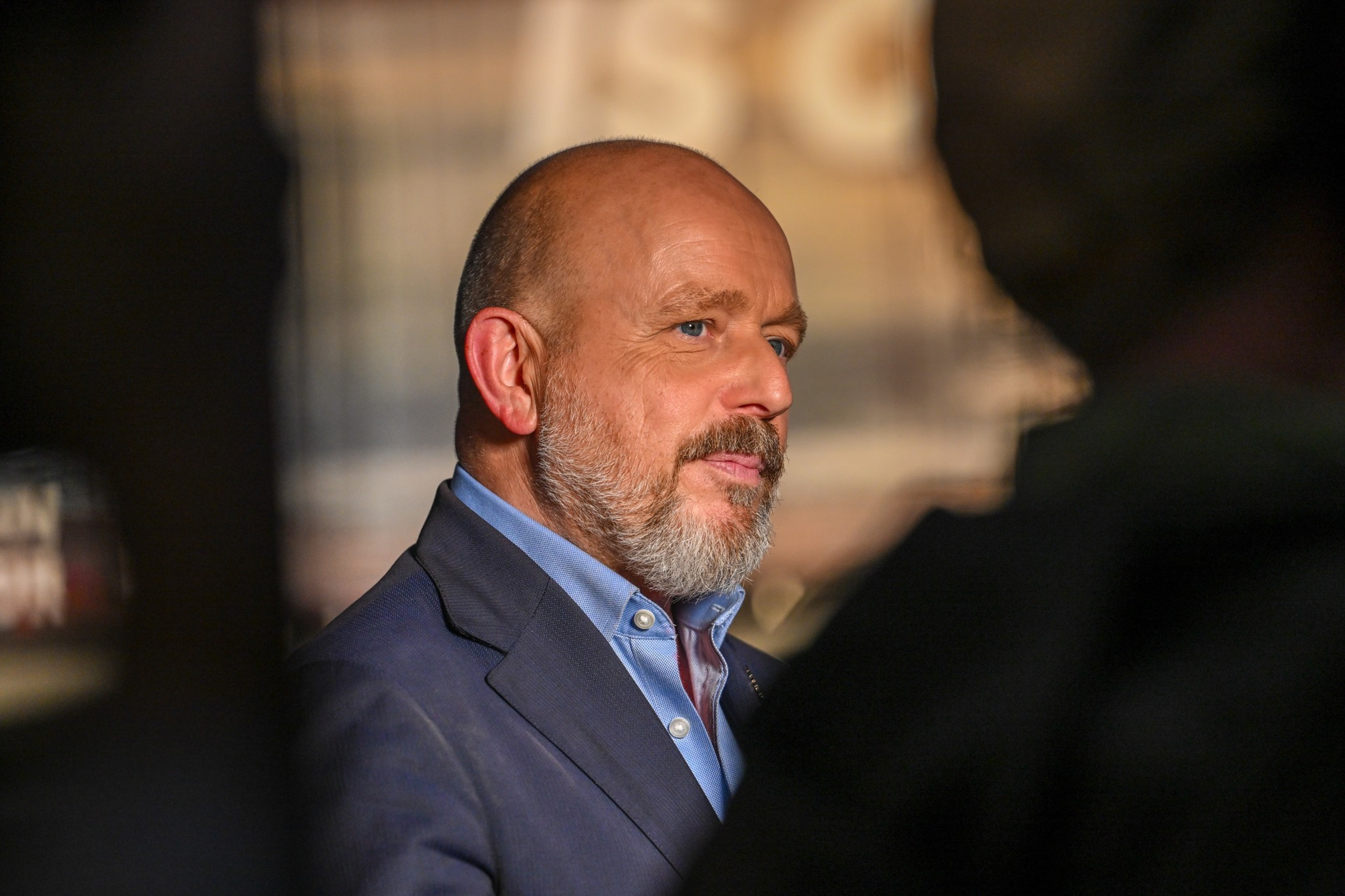 A middle-aged man with a bald head and gray beard wears a blue dress shirt and dark blazer, looking attentively to his right.