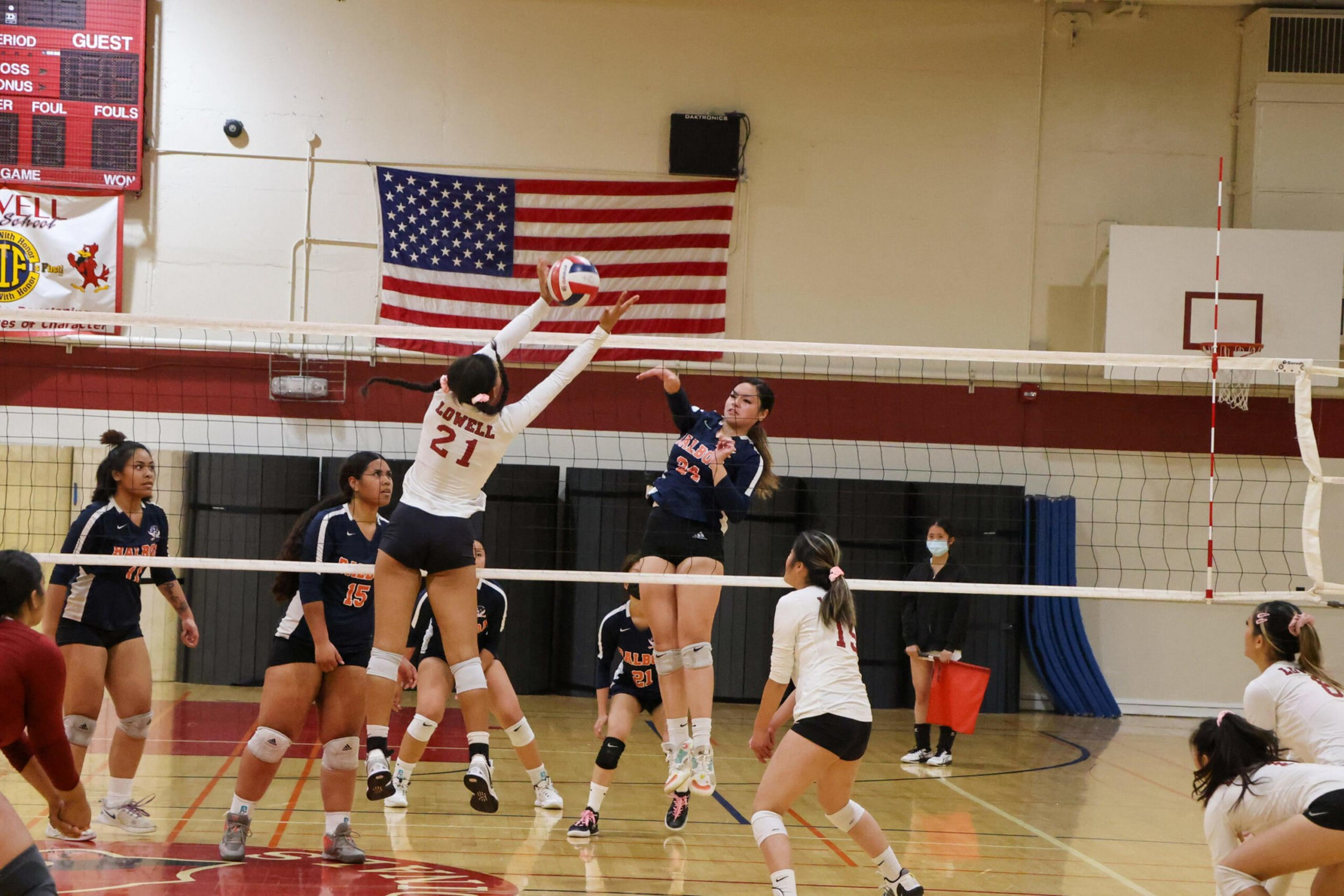 Two volleyball players jump near the net, one blocking and one spiking the ball, while teammates watch in a gym with a U.S. flag on the wall.