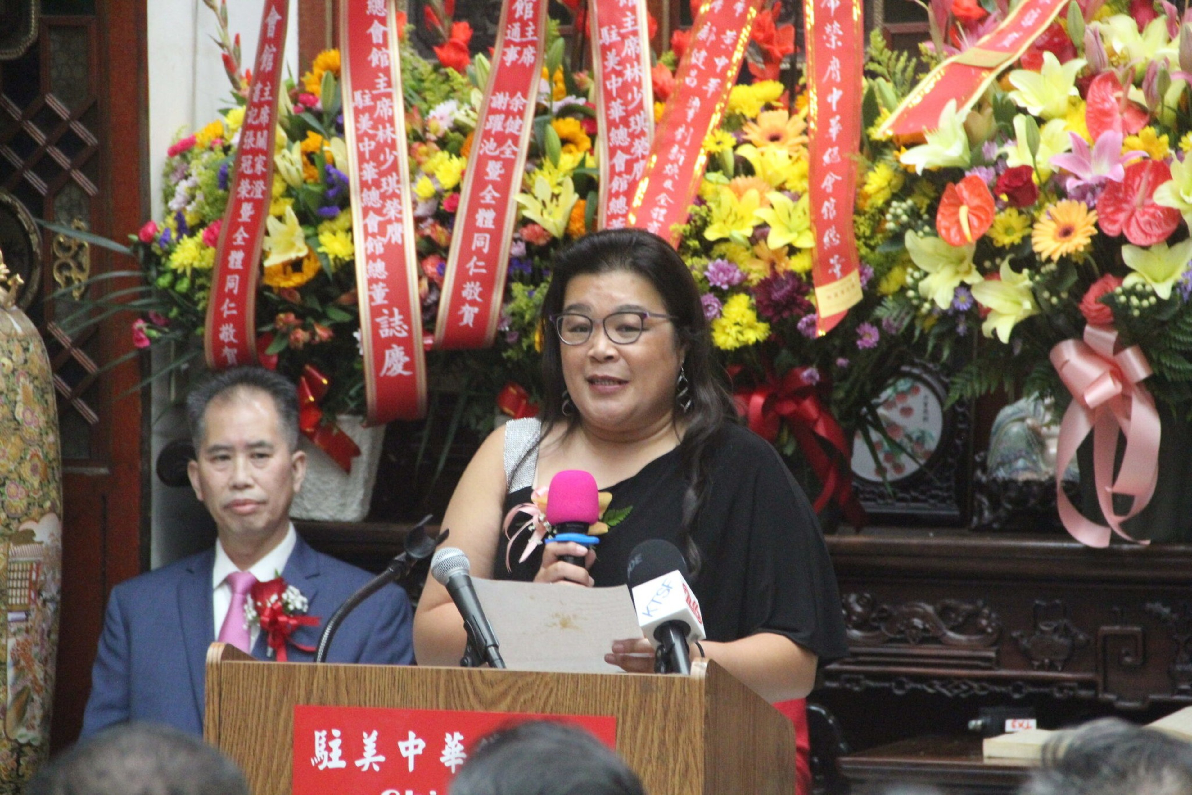 A woman with glasses speaks into a pink microphone at a podium, with a man in a suit seated beside her and colorful floral arrangements behind them.