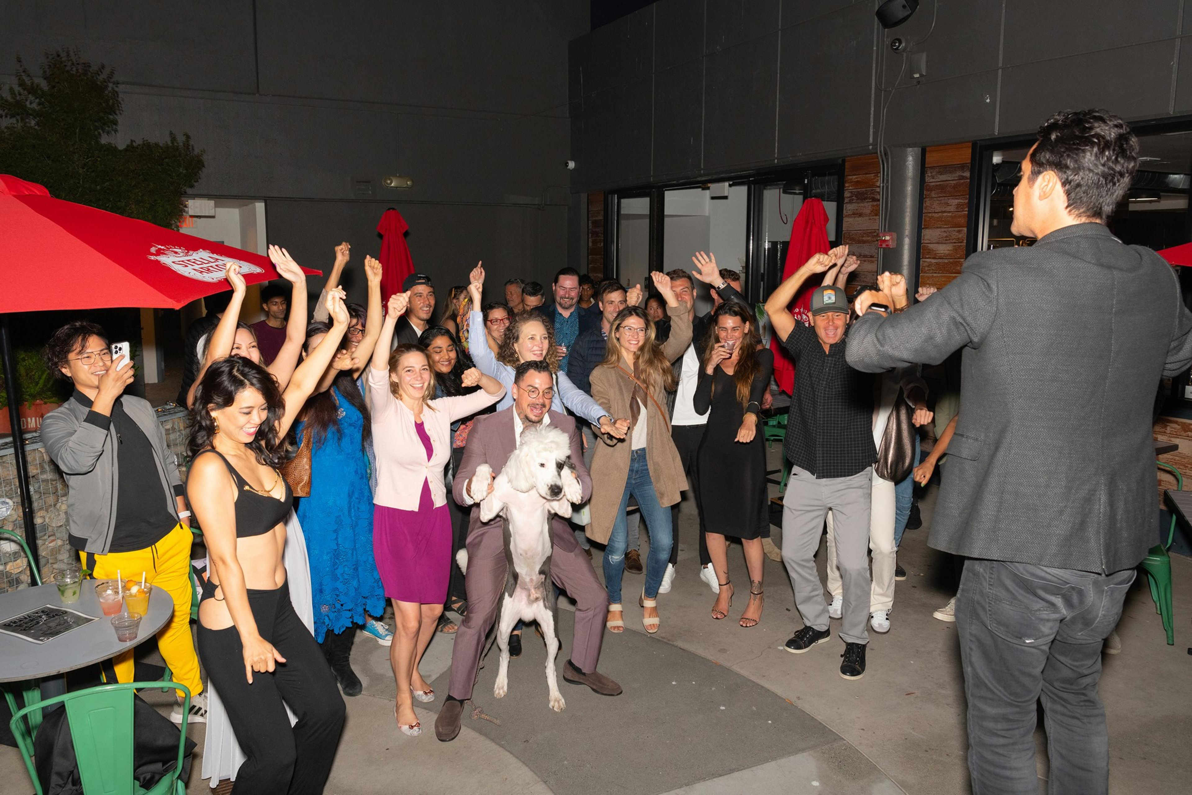 A group of people is joyfully dancing at an outdoor party, with vibrant expressions and dynamic poses. A man is holding a dog, and red umbrellas are in the background.