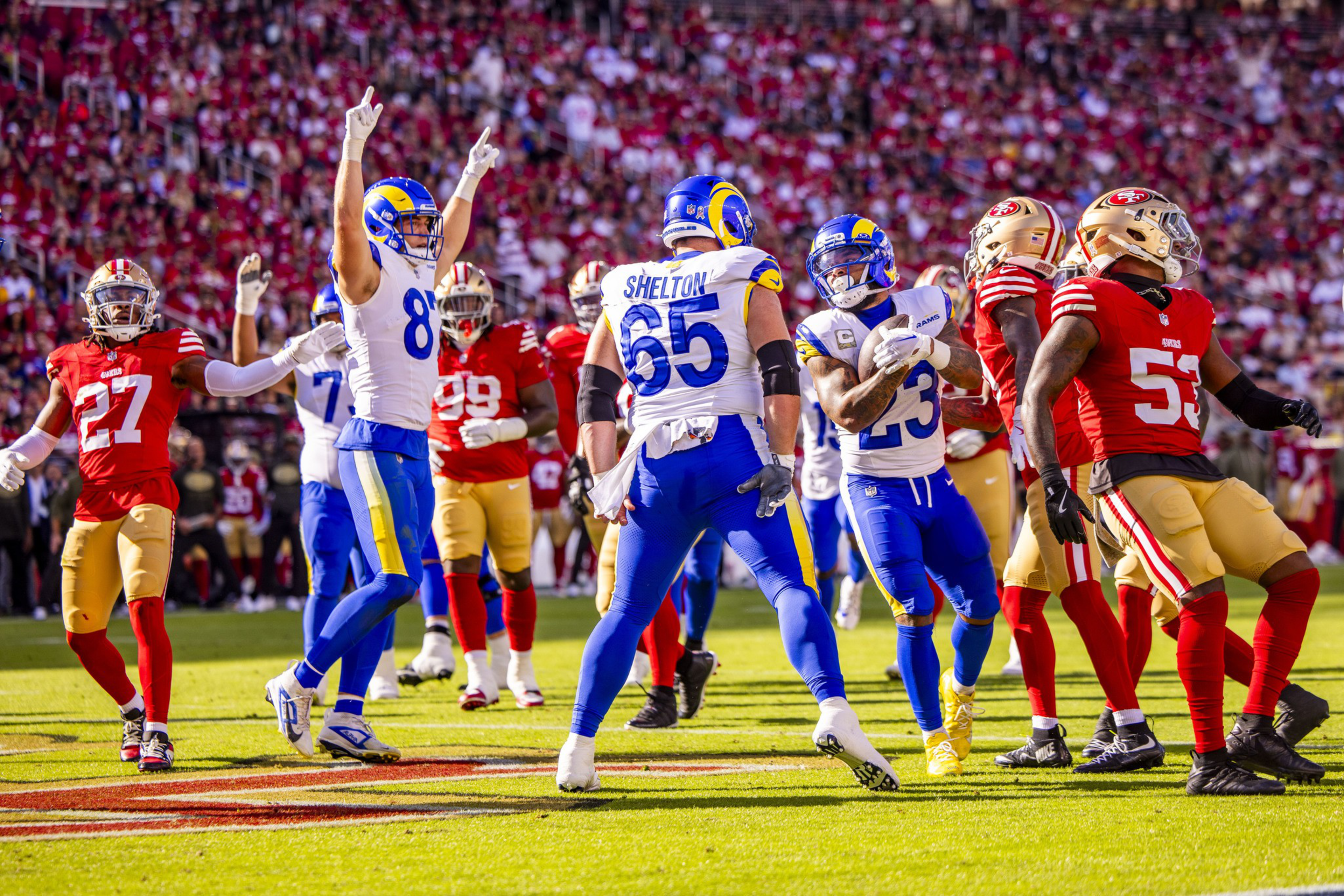 Players in blue and white celebrate on the field while players in red and gold stand nearby during a football game with a packed crowd in the background.