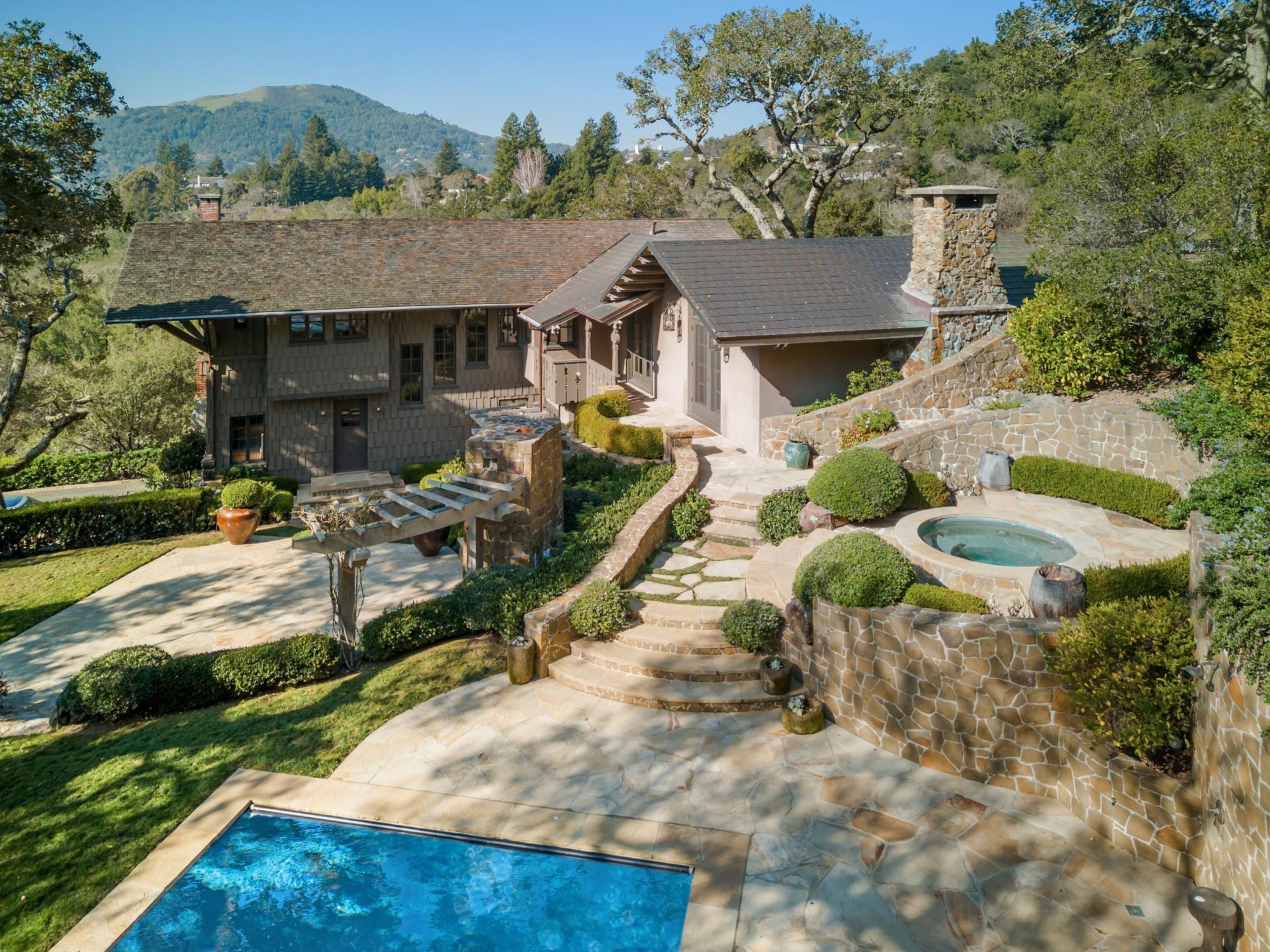 A two-story house with a stone patio, a pool, a round hot tub surrounded by stone walls, greenery, and a mountainous landscape in the background.