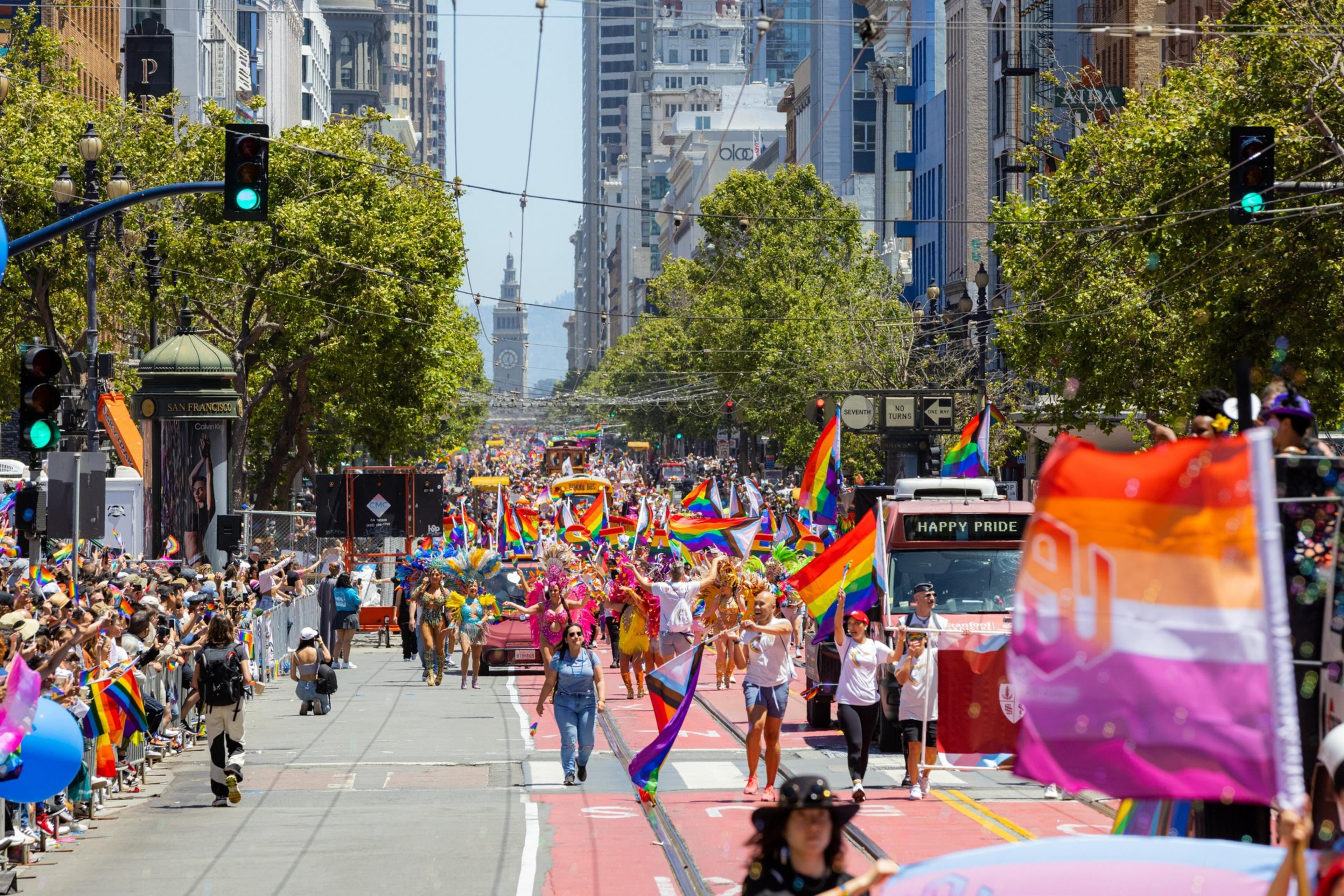A vibrant pride parade with people carrying rainbow flags, colorful outfits, and enthusiastic crowds lining a sunlit city street.