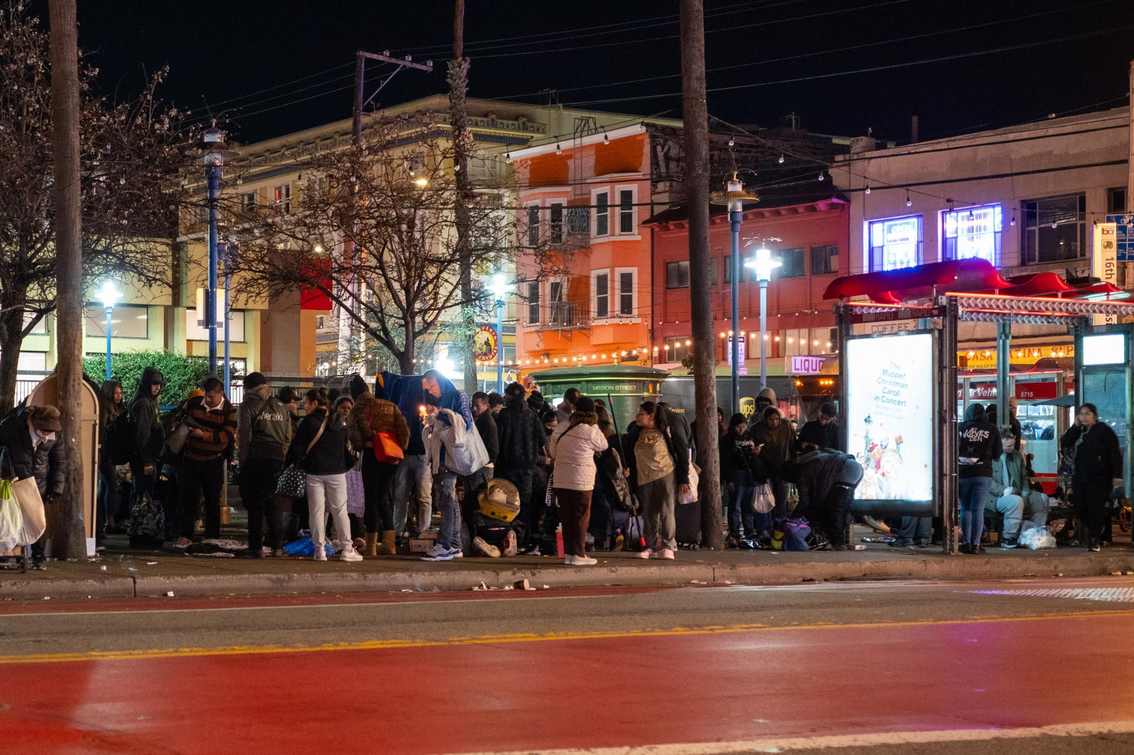 A group of people gather at a bus stop on a city street at night. Brightly lit buildings are in the background, and the scene is bustling with activity.