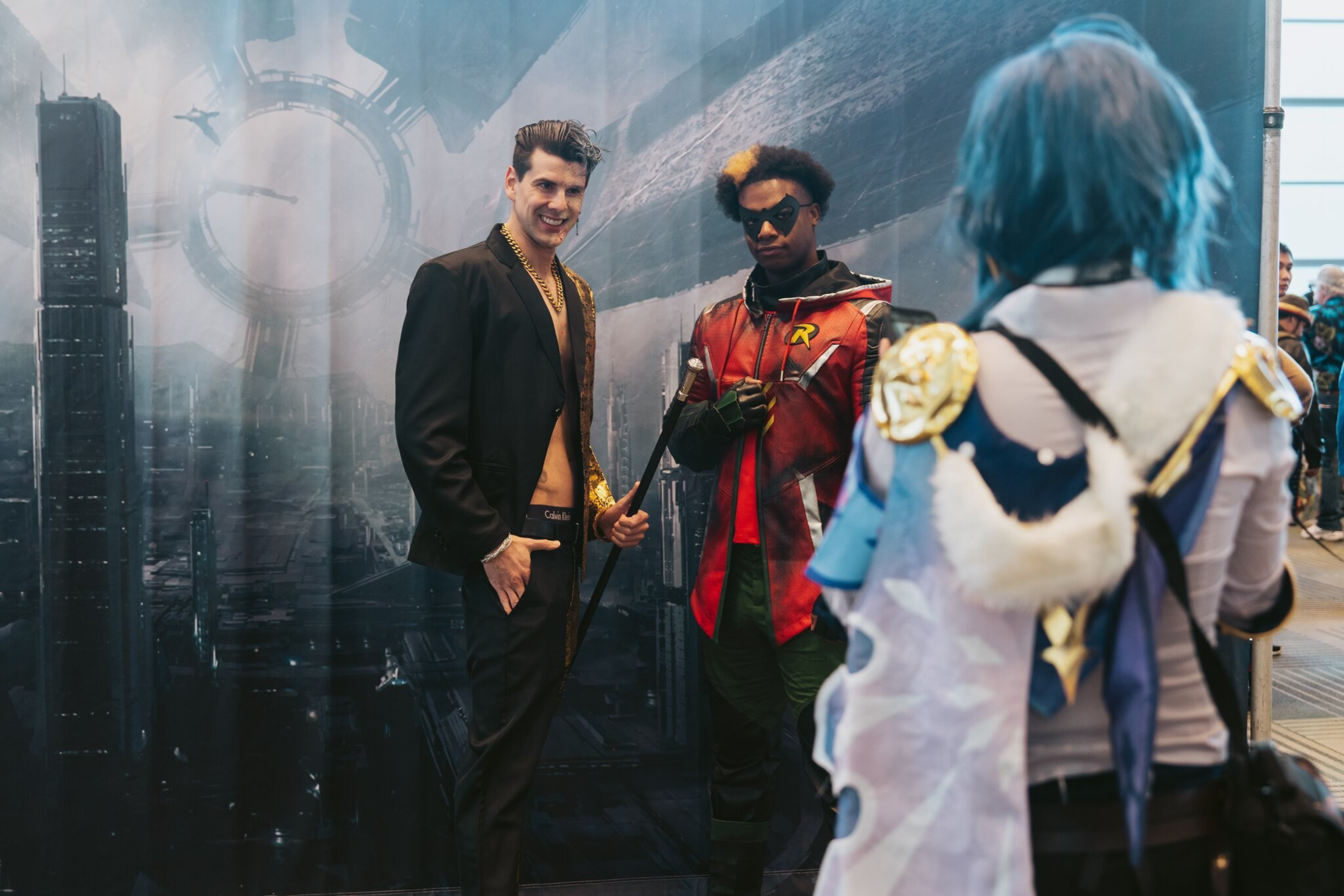 Two men in dashing costumes stand in front of a wall made to look like a sky.