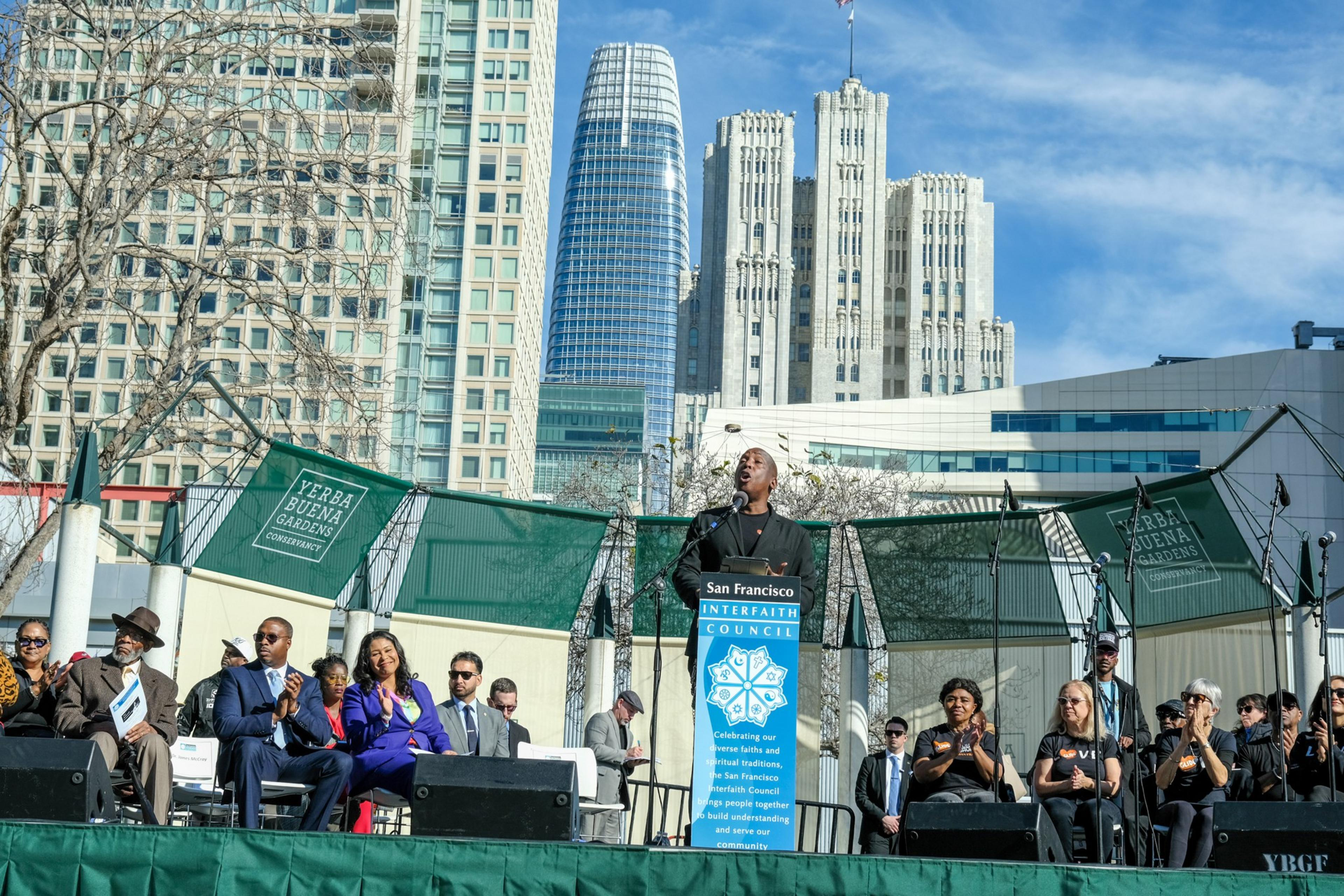 A person speaks at a podium on a stage, with a city skyline in the background and an attentive audience seated in front.