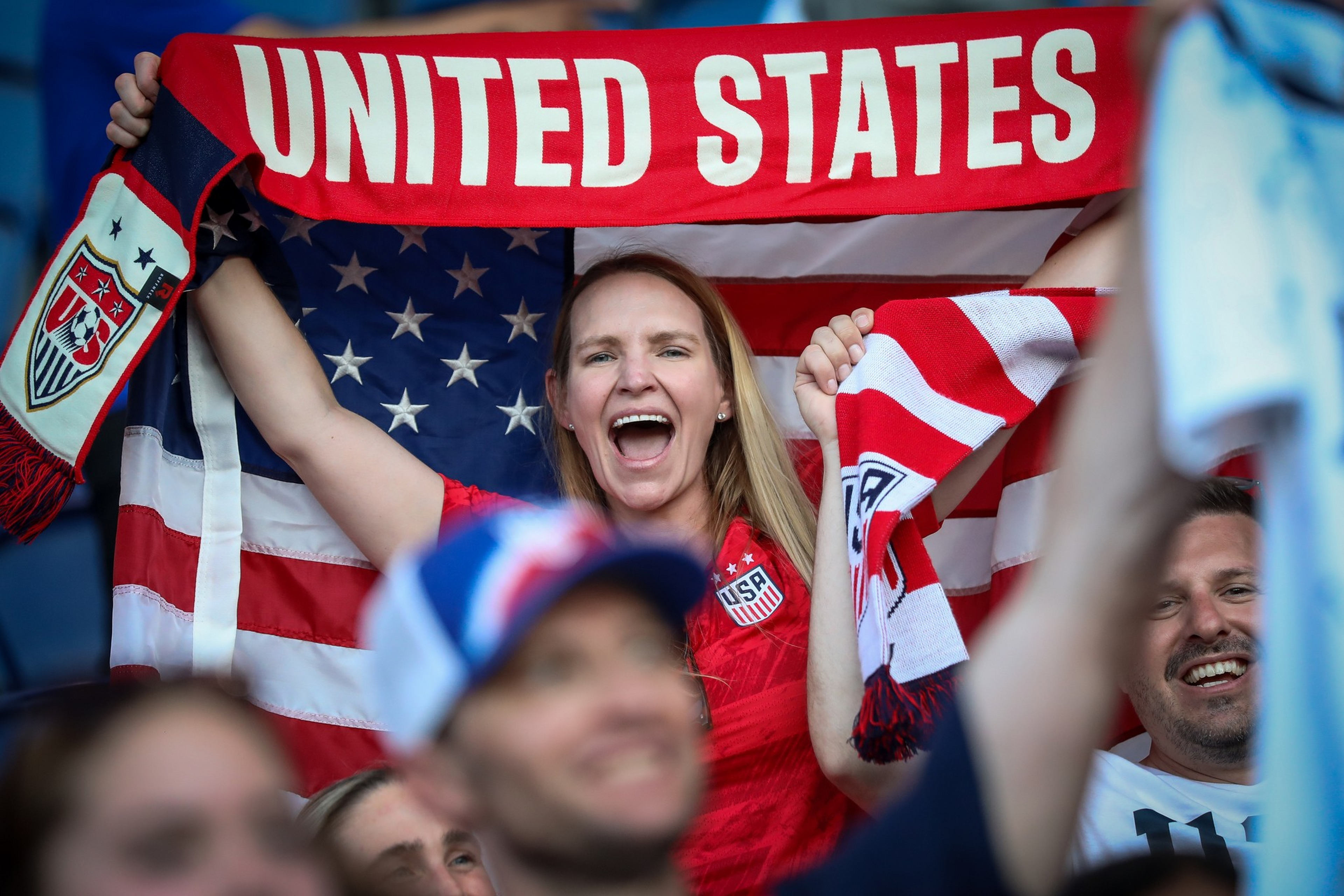 A smiling woman holds up a red scarf reading “UNITED STATES” with the American flag behind her and fans around her.