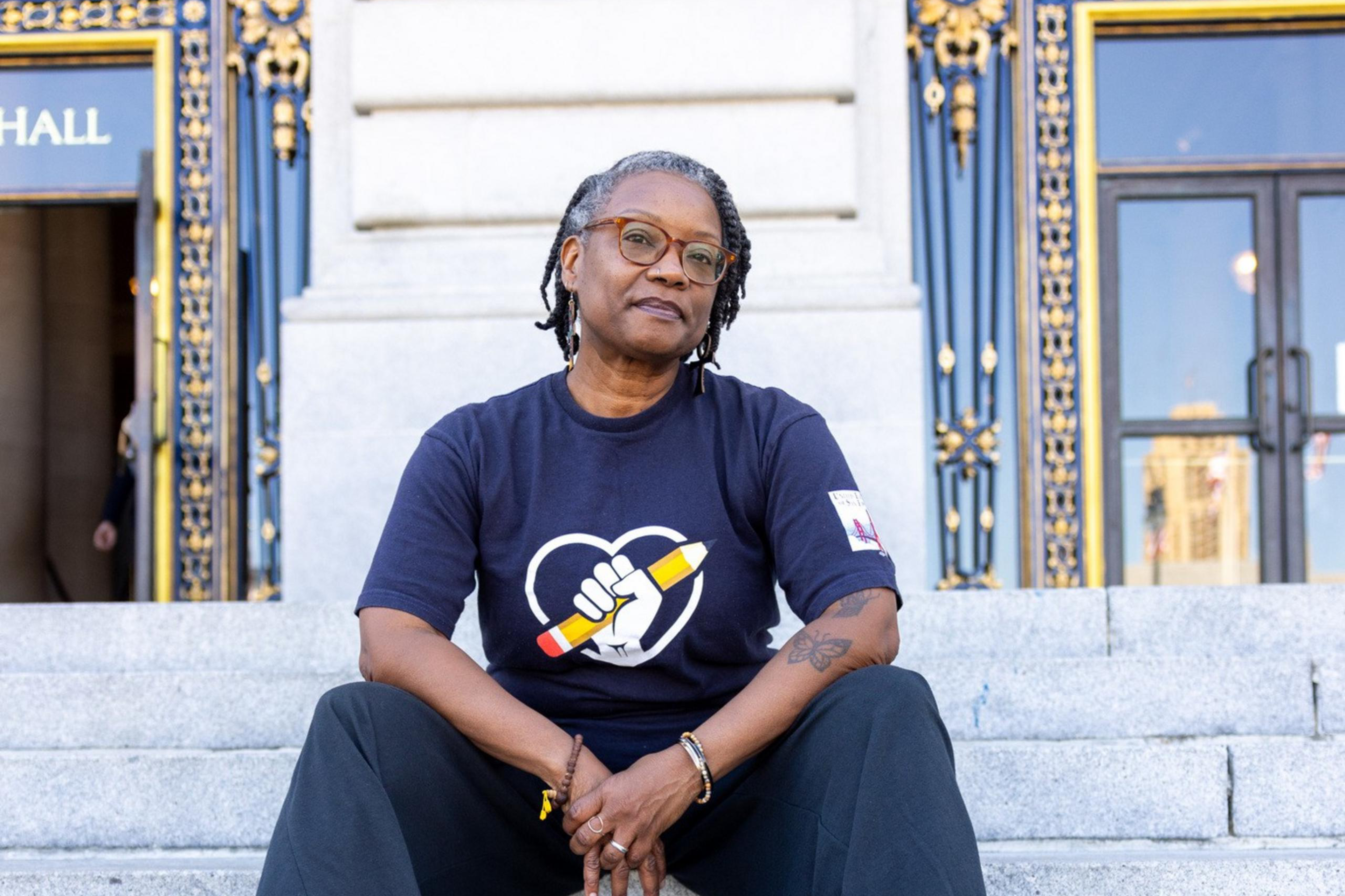 A person with dreadlocks, wearing glasses, a navy shirt with a fist and pencil logo, dark pants, and black Crocs, sits on ornate building steps with golden details.