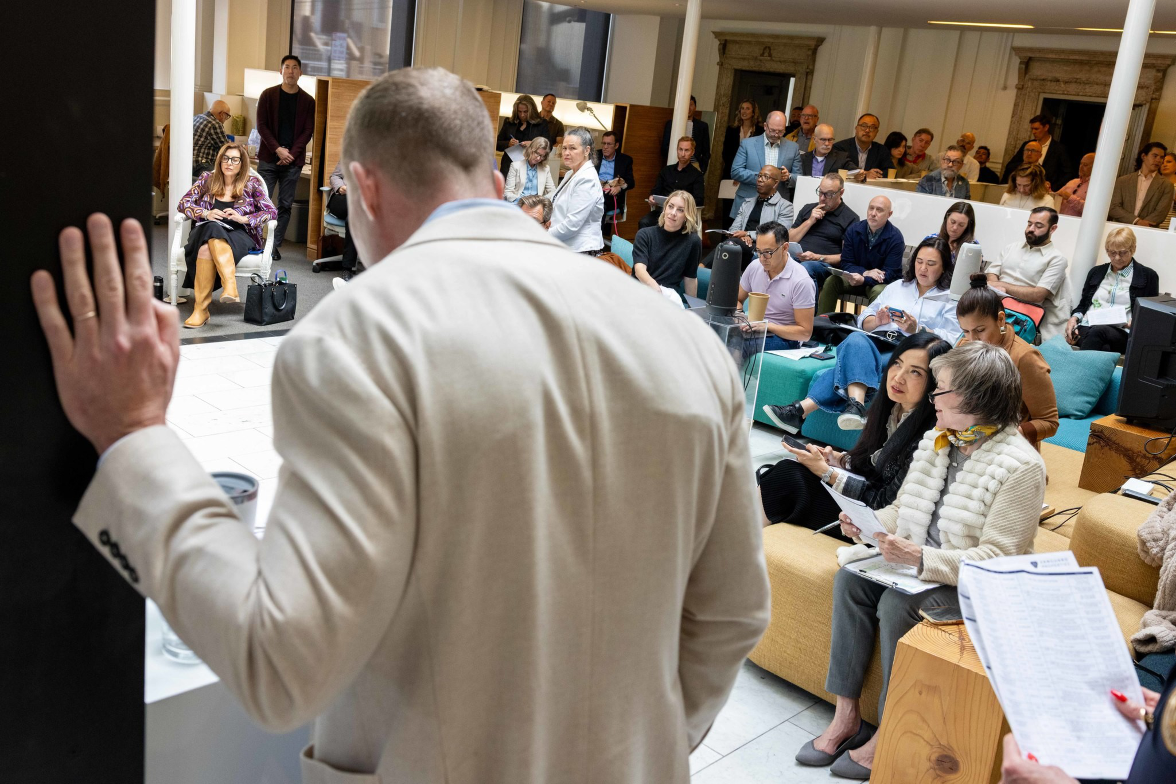 A man in a beige jacket stands addressing a seated audience of diverse adults, some taking notes or looking attentively in a large, bright room.