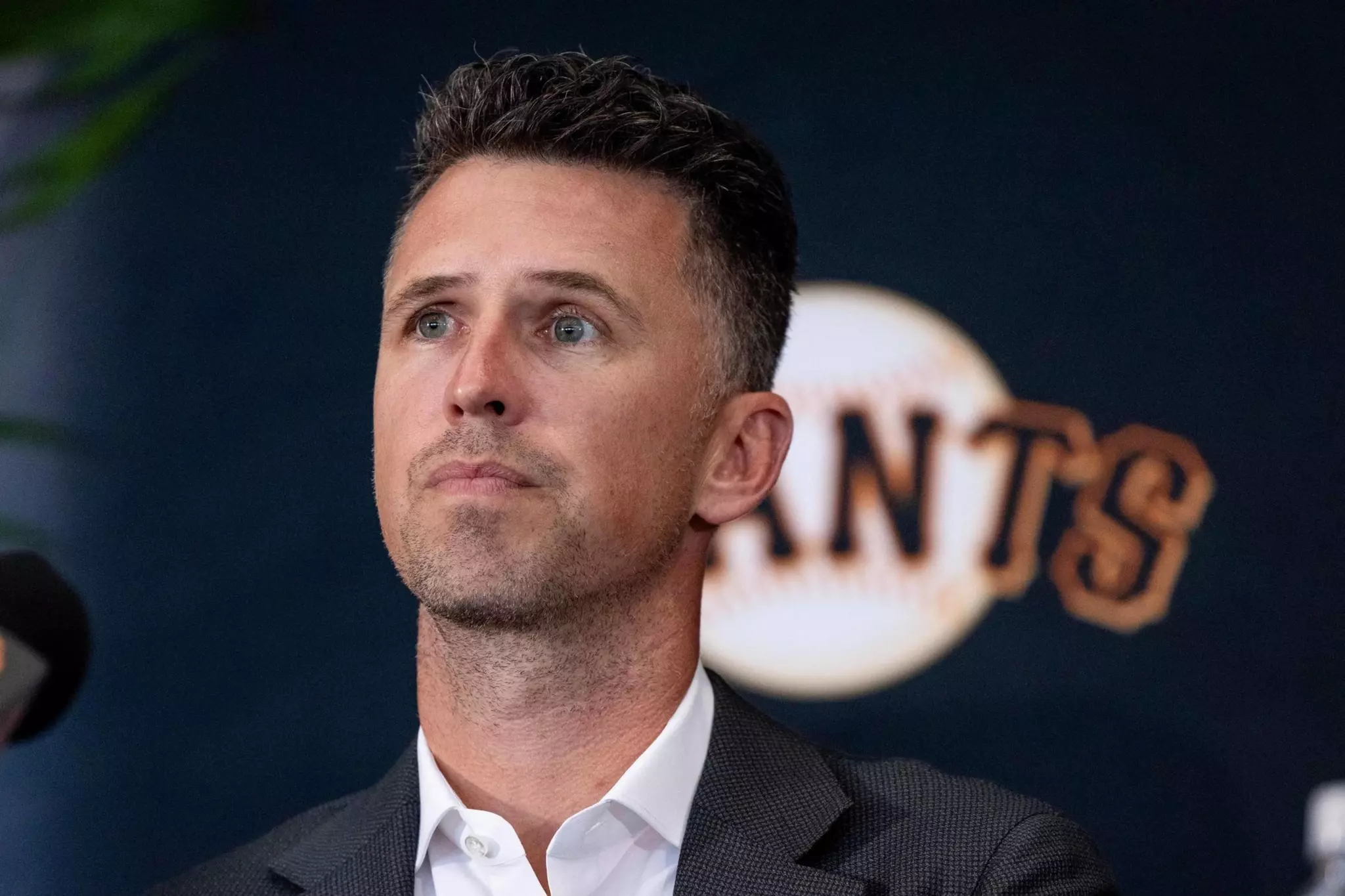 A person with short hair is wearing a suit and looking thoughtful. Behind them is a wall with a design resembling a baseball and the word "GIANTS."