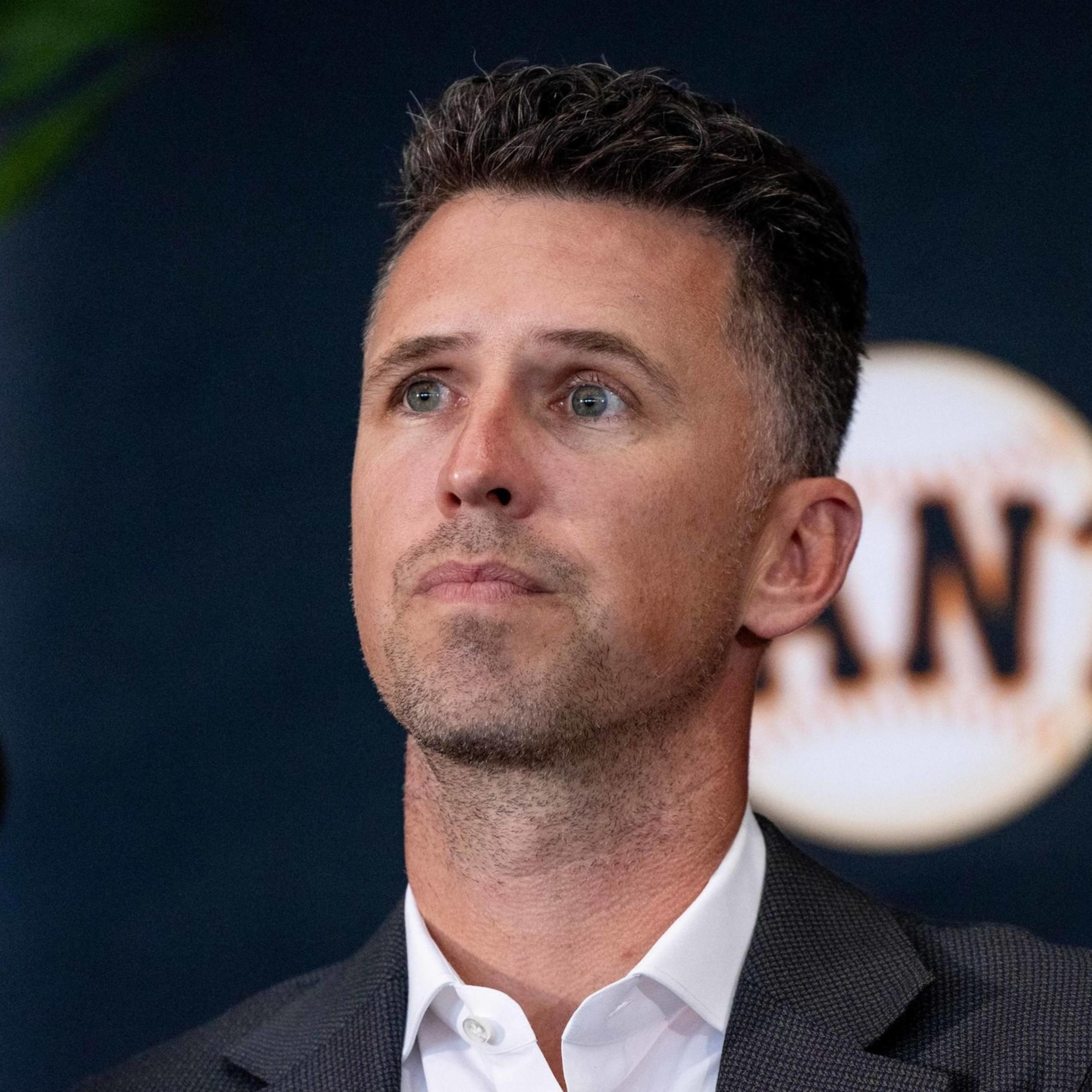 A person with short hair is wearing a suit and looking thoughtful. Behind them is a wall with a design resembling a baseball and the word "GIANTS."