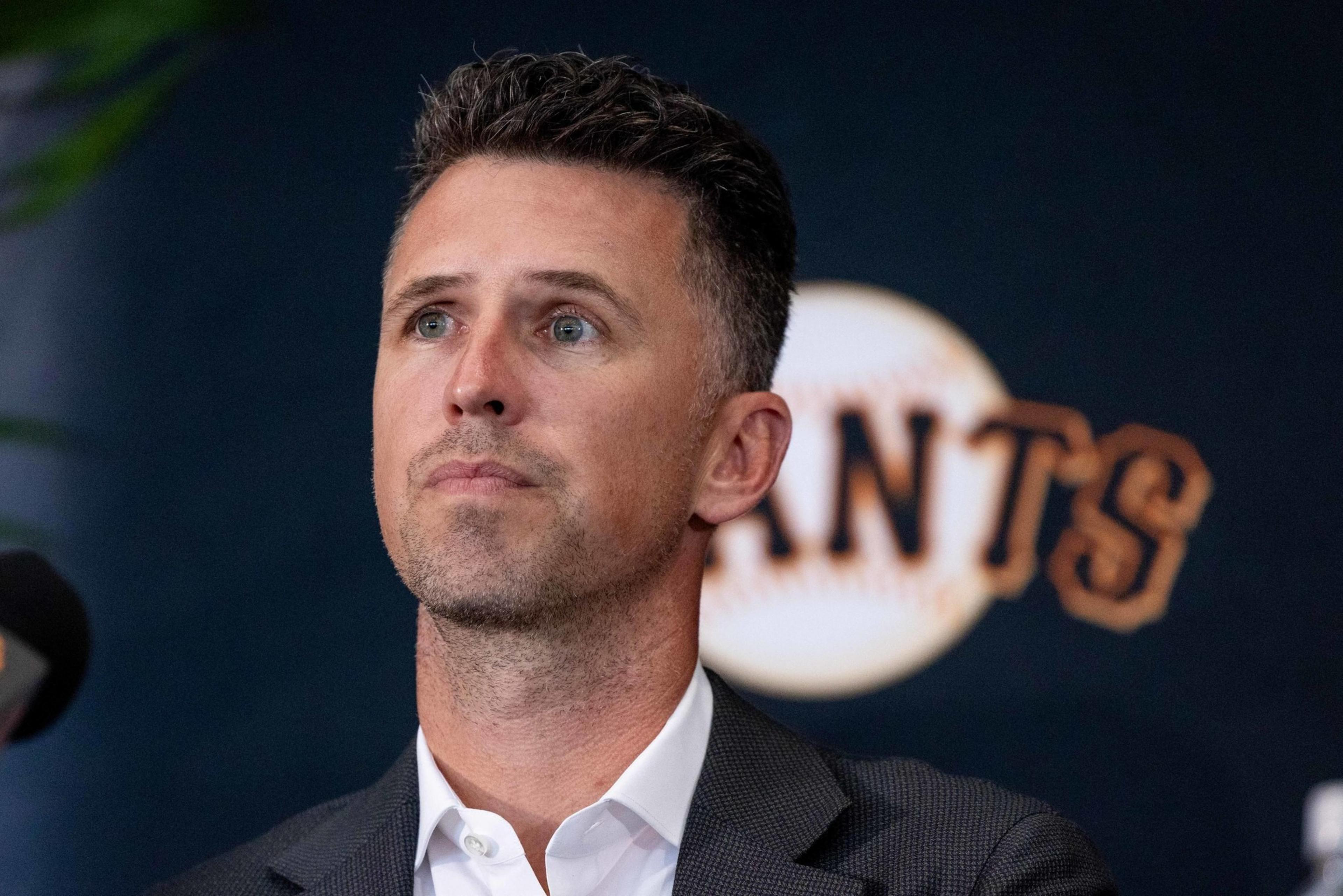 A person with short hair is wearing a suit and looking thoughtful. Behind them is a wall with a design resembling a baseball and the word "GIANTS."