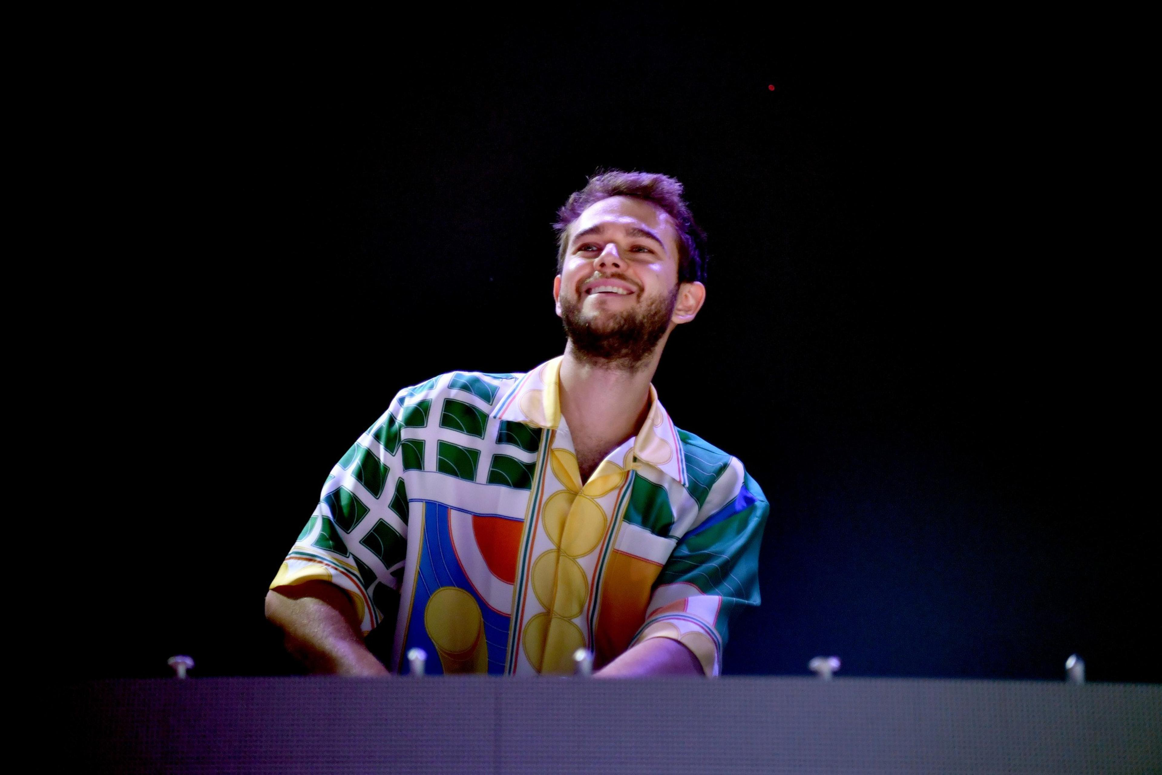 A man with short hair and a beard smiles while standing at a DJ booth. He's wearing a colorful geometric-patterned shirt against a dark background.
