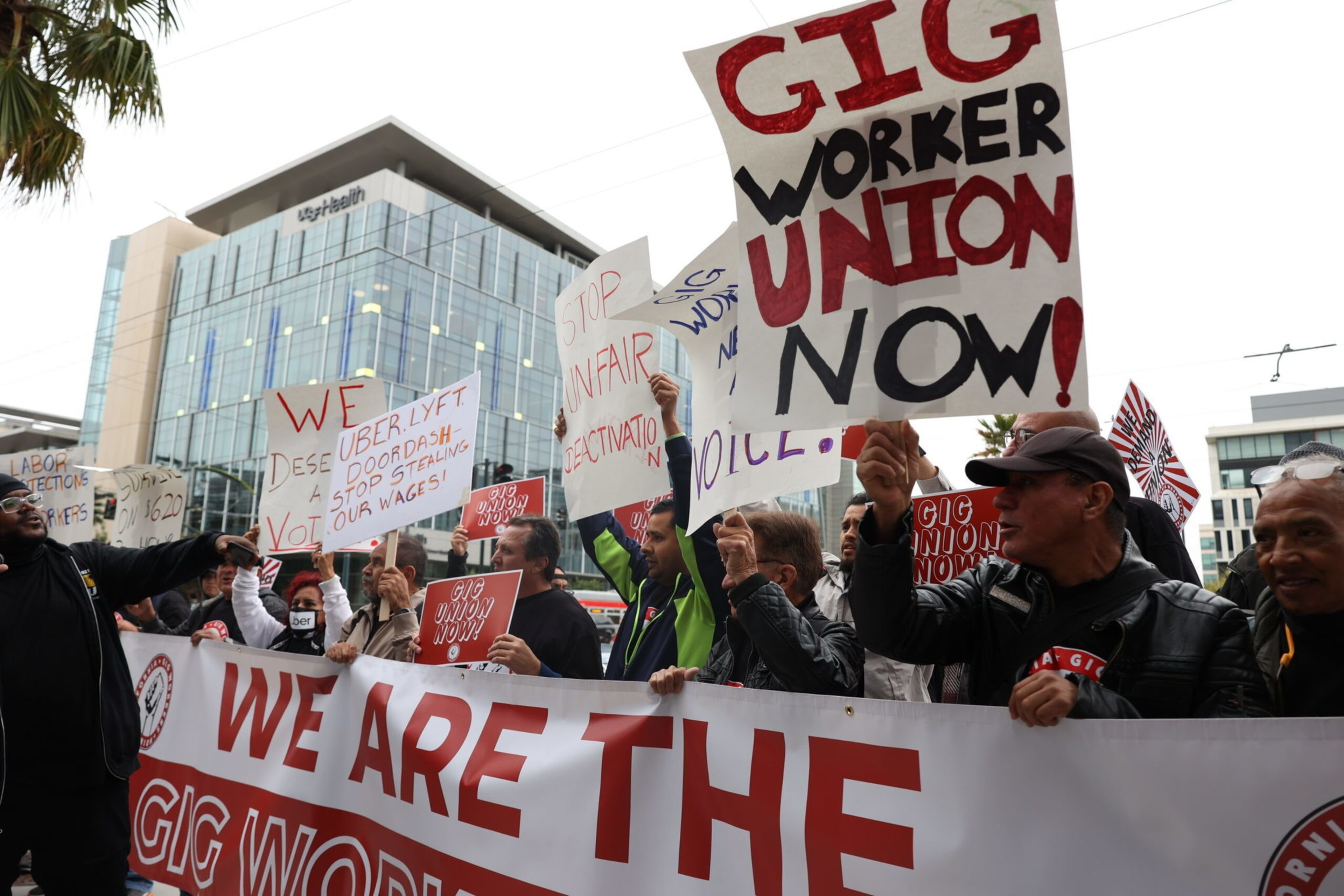 People hold signs demanding a gig worker union and fair wages, protesting outside a large building with glass windows.