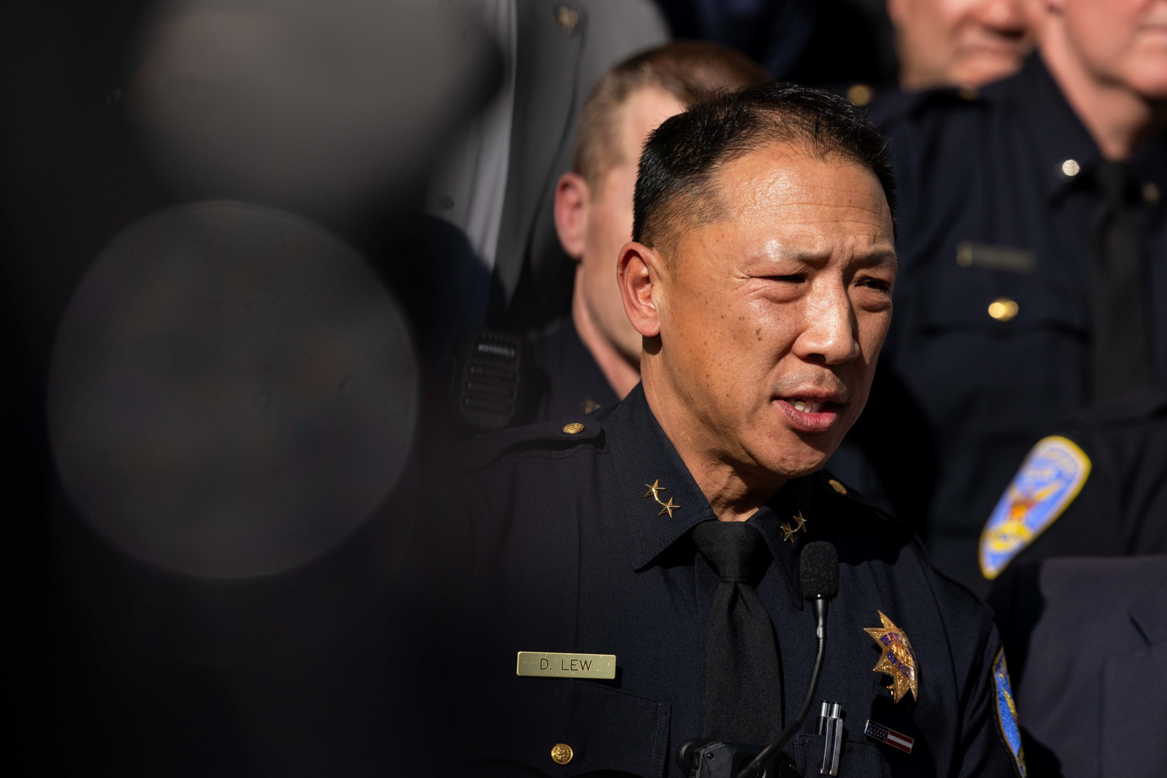 A police officer with the name tag "D Lew" speaks into a microphone, wearing a dark uniform with gold stars on the collar and a badge on the chest.
