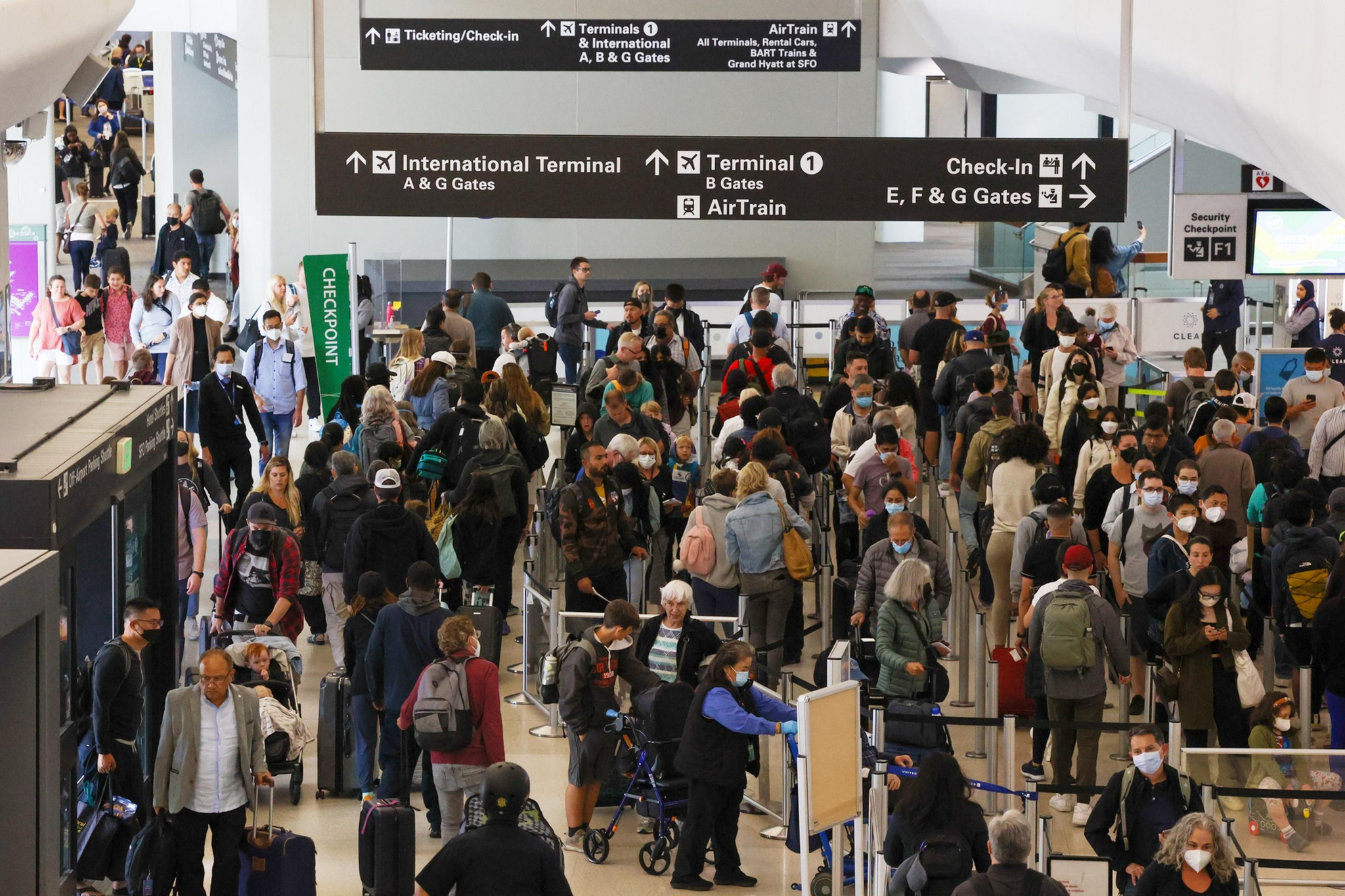 A crowded airport terminal features travelers wearing masks, many with luggage, standing in long lines at a security checkpoint under signs directing to terminals and gates.
