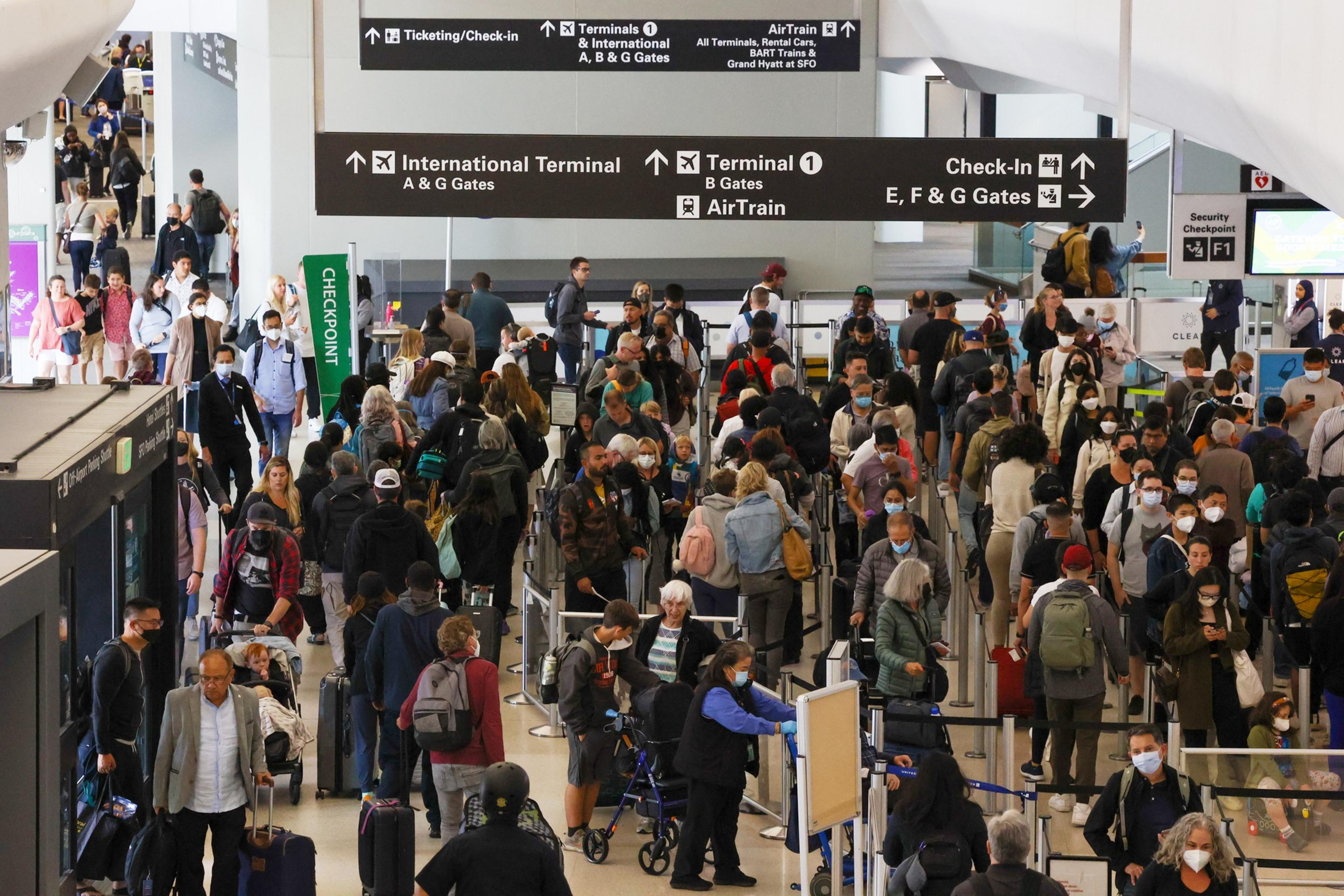 A crowded airport terminal features travelers wearing masks, many with luggage, standing in long lines at a security checkpoint under signs directing to terminals and gates.