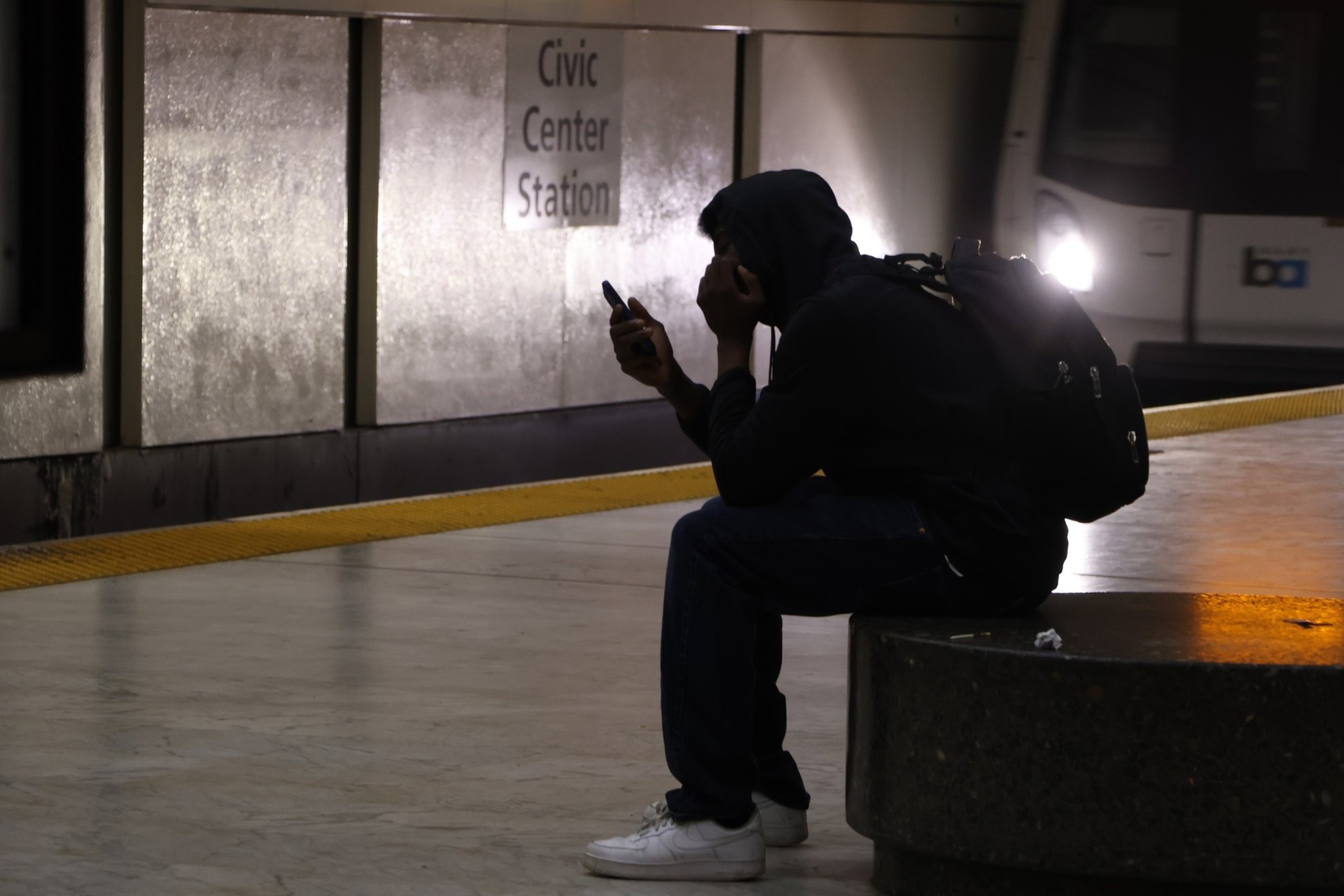 A person wearing a hoodie and backpack sits on a bench at Civic Center Station, looking at their phone with a train approaching.