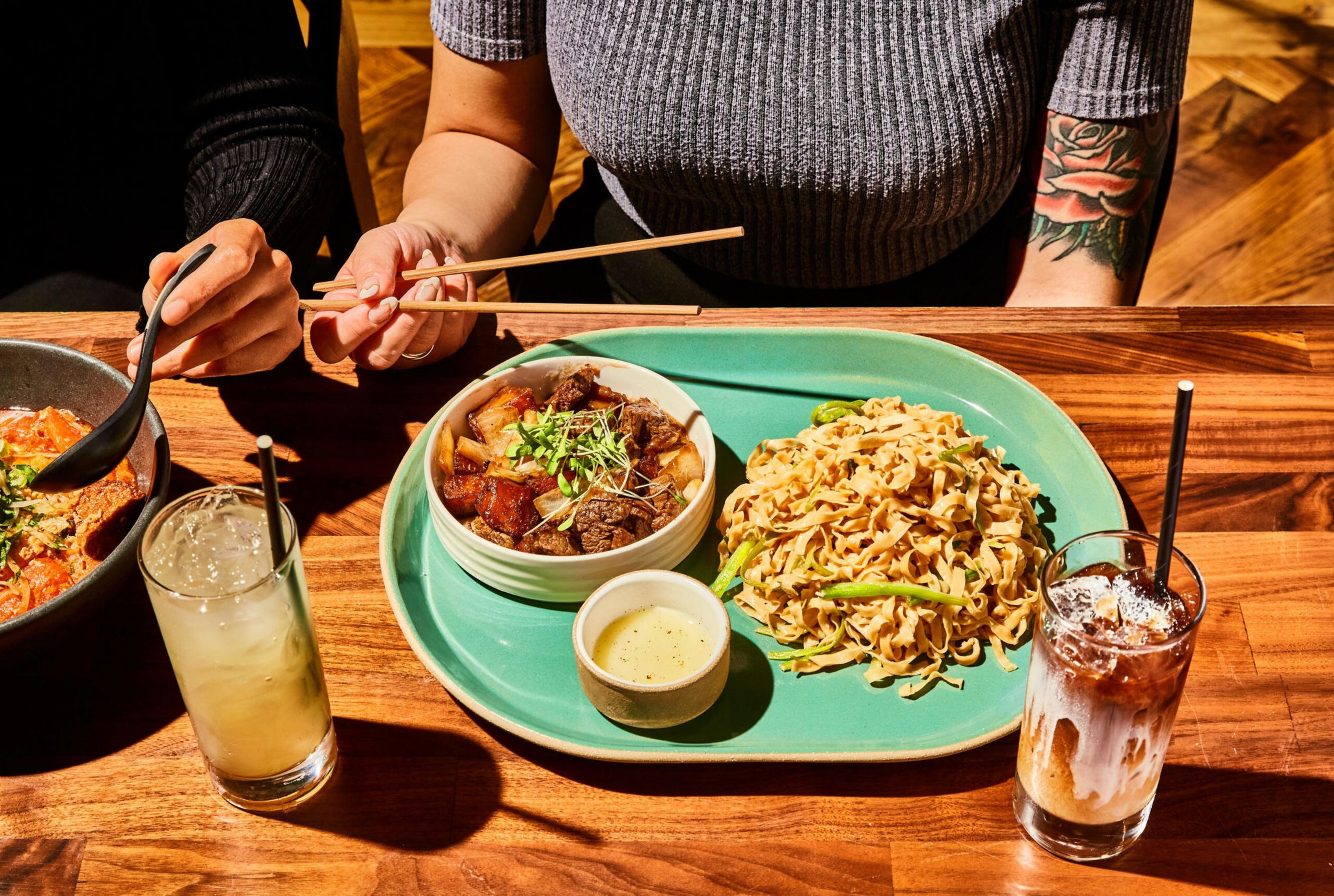 Two people are dining with chopsticks, enjoying a meal of noodles and beef stew. Drinks sit nearby on a polished wooden table.