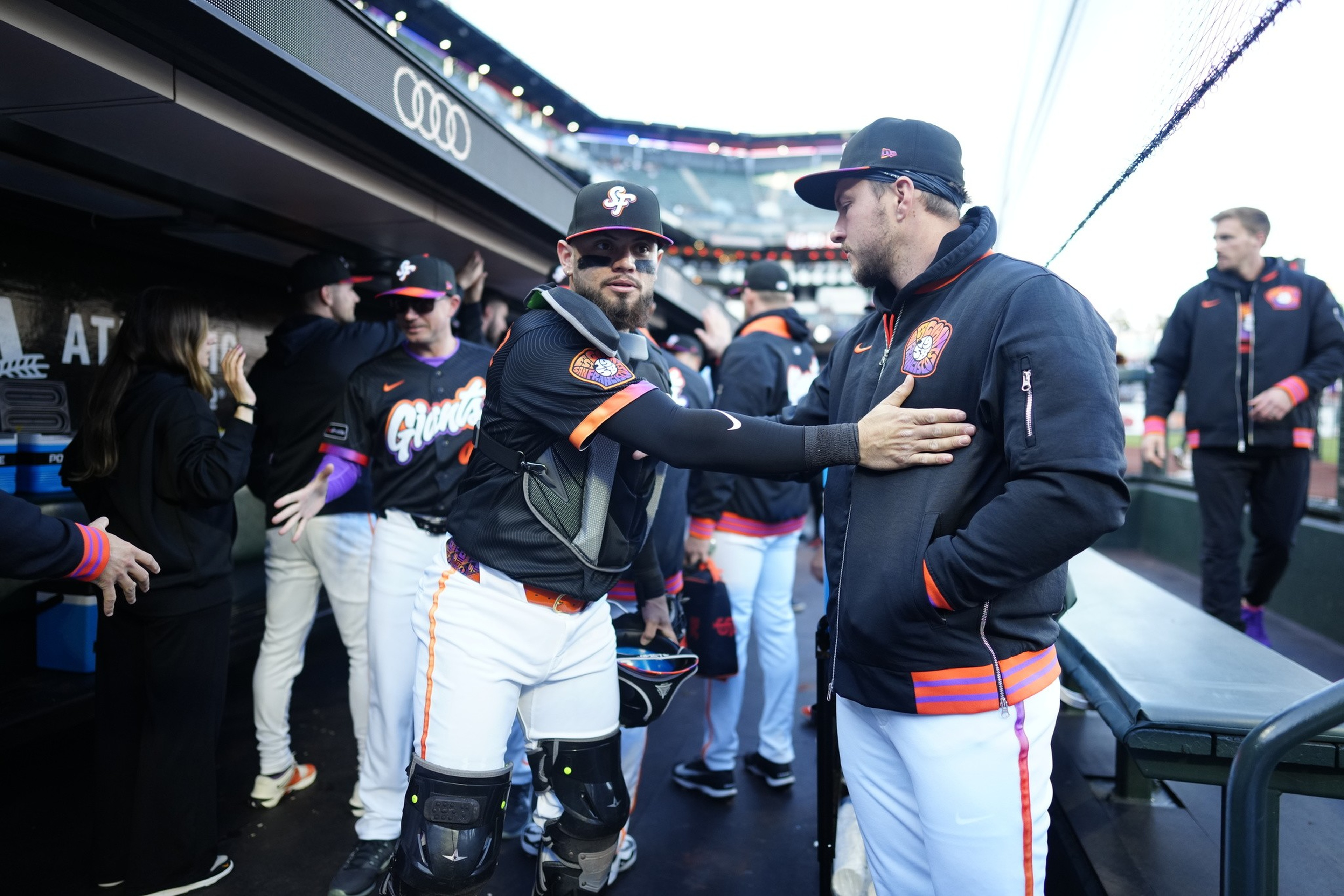 Baseball players in black and orange uniforms interact in a dugout, with one player reaching out to touch another’s chest.