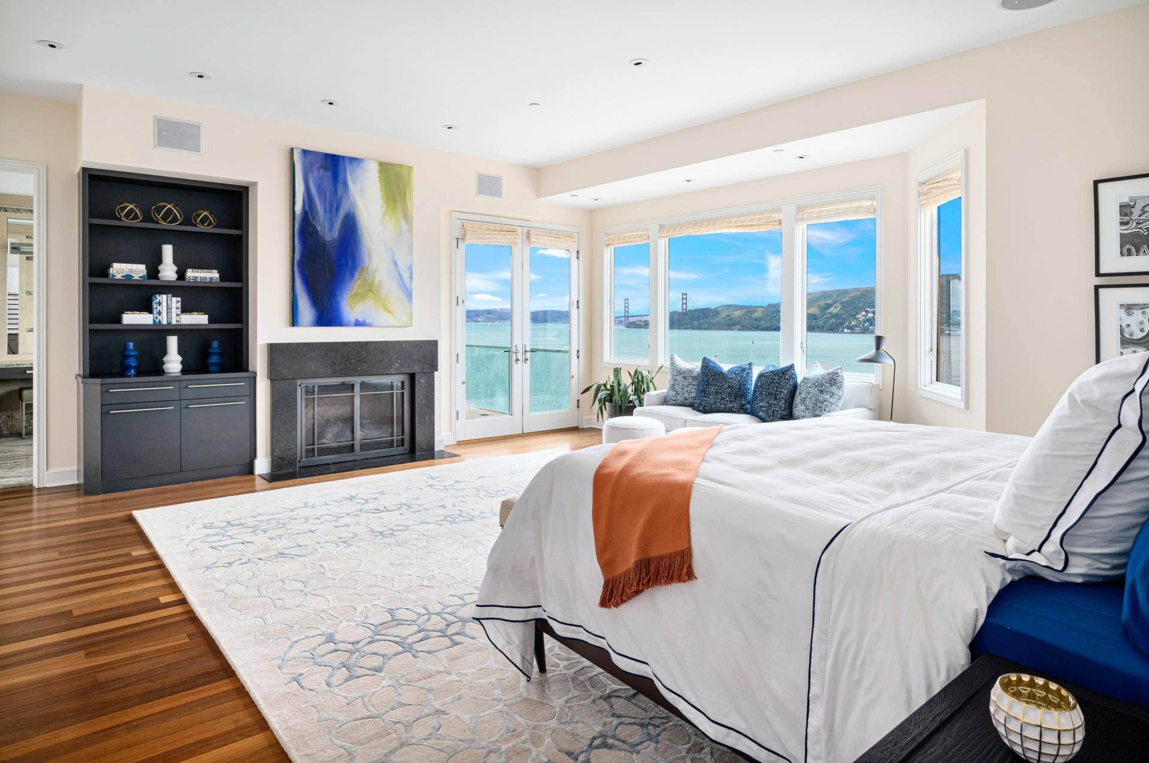 A spacious bedroom features a large bed with white bedding and an orange throw, hardwood floors, a fireplace, built-in shelves, and windows overlooking water and a bridge.