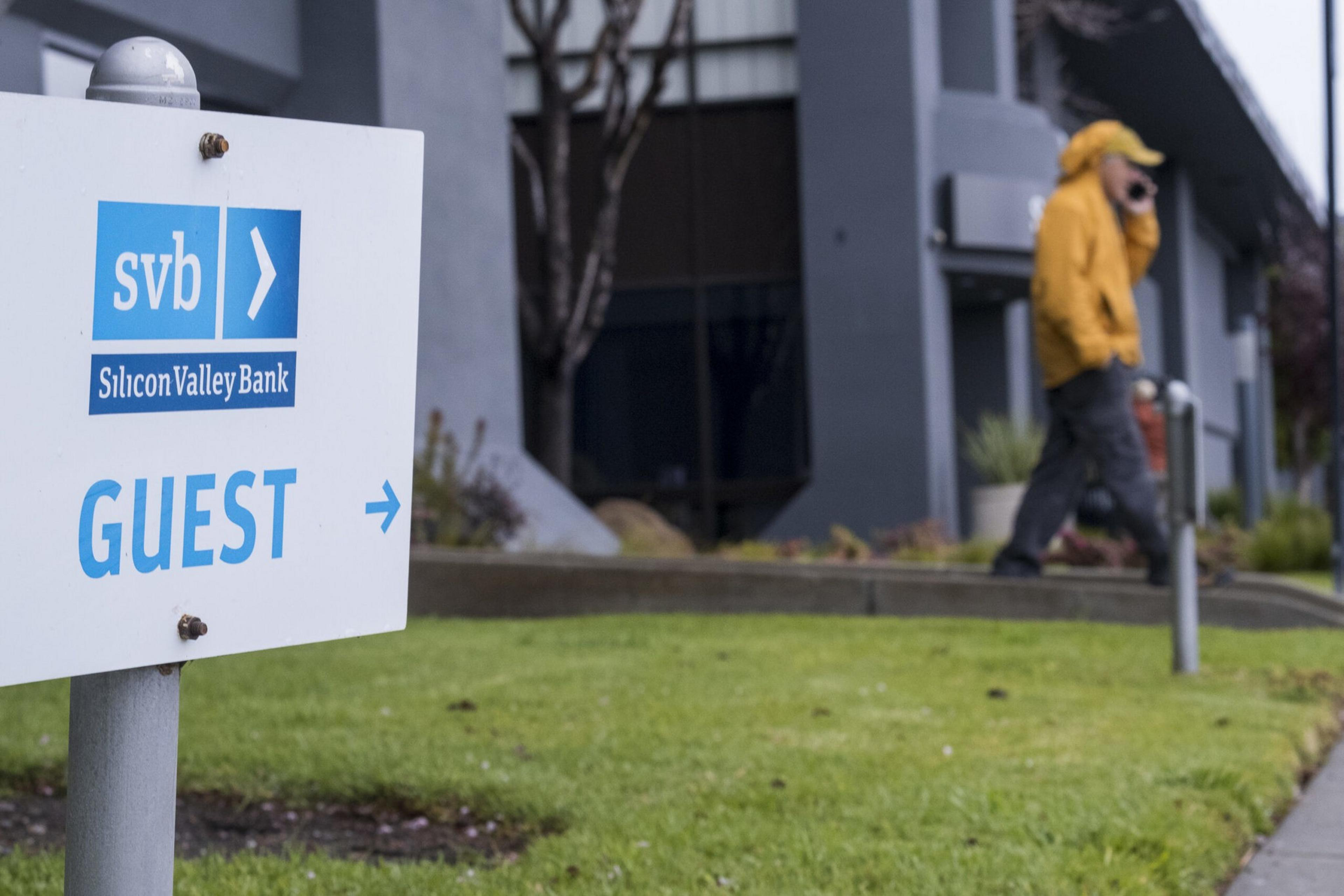 A Silicon Valley Bank guest parking sign is in the foreground with a person in a yellow jacket walking and talking on a phone near a building in the background.