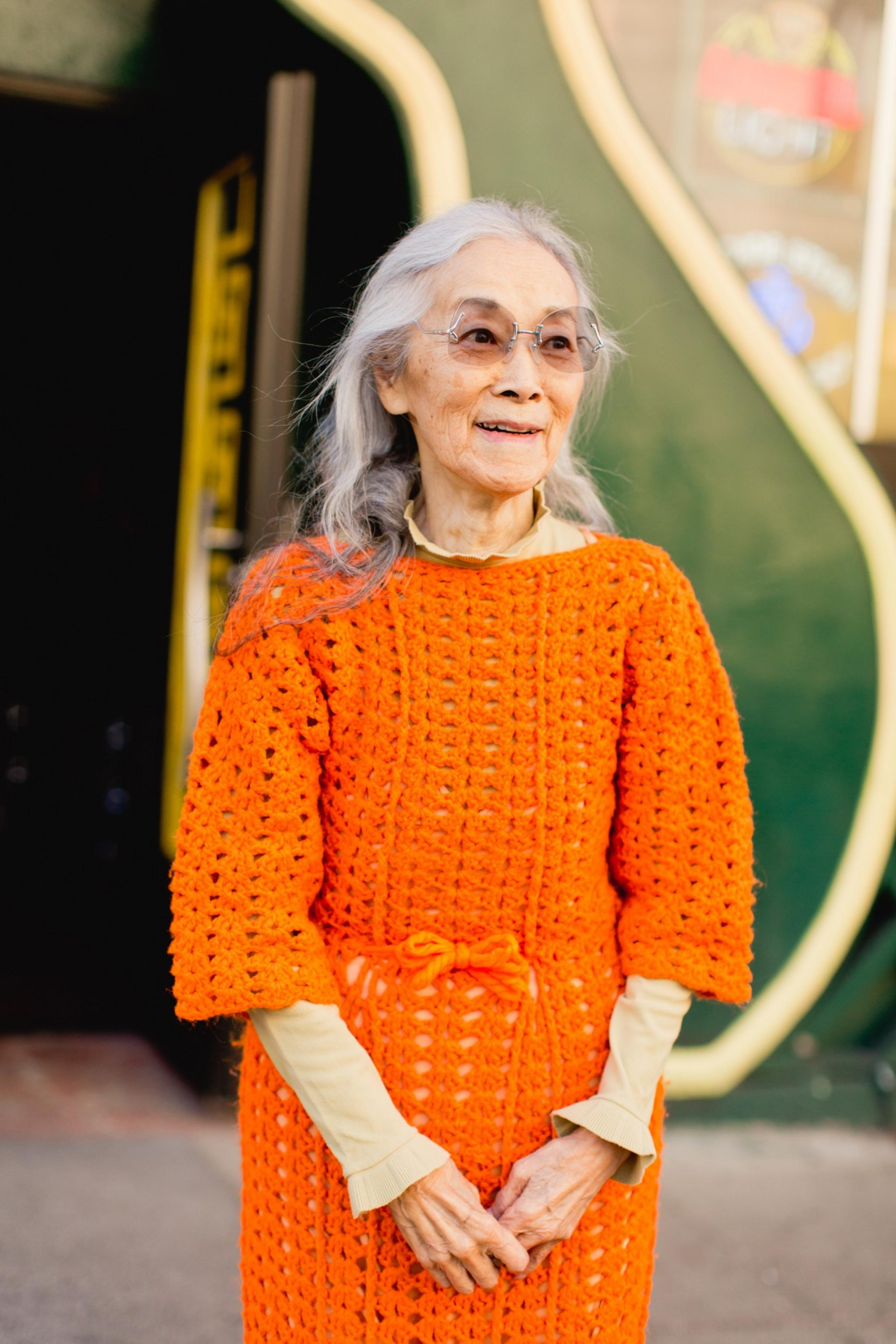An elderly woman with gray hair and glasses smiles while wearing a bright orange crocheted dress over a light yellow shirt.