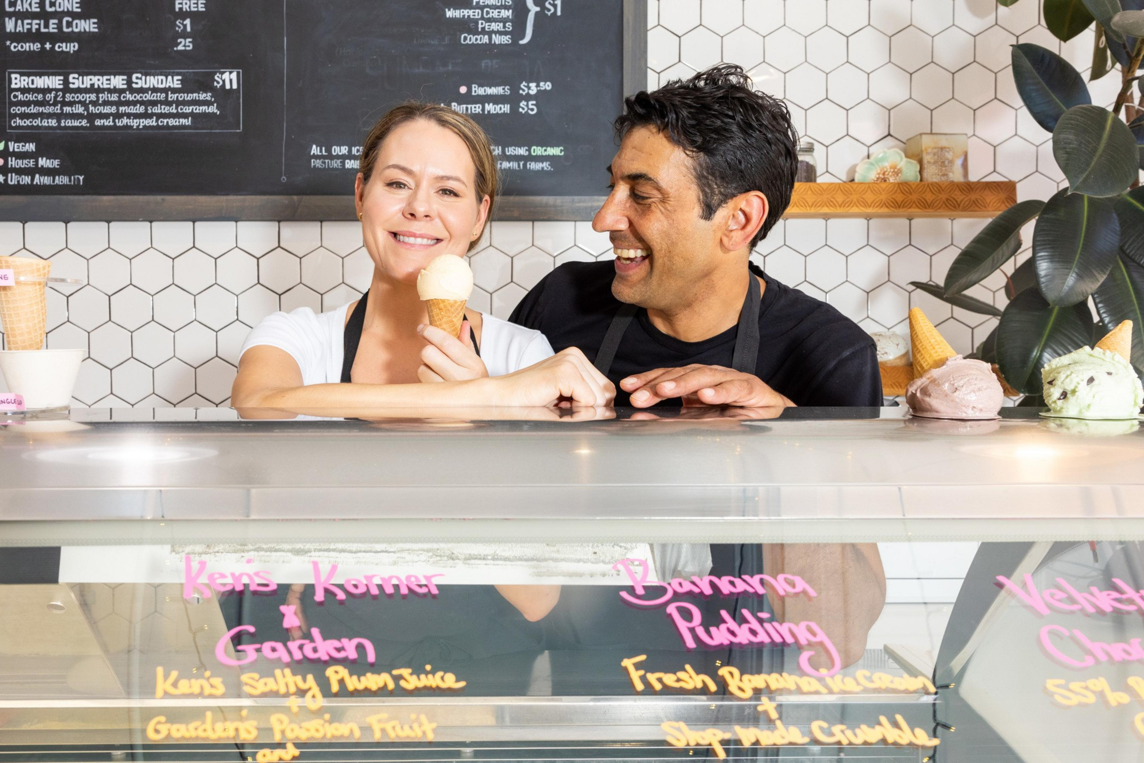 Two people behind an ice cream counter smile as the woman holds an ice cream cone. The menu on the wall behind them lists various ice cream flavors and prices.