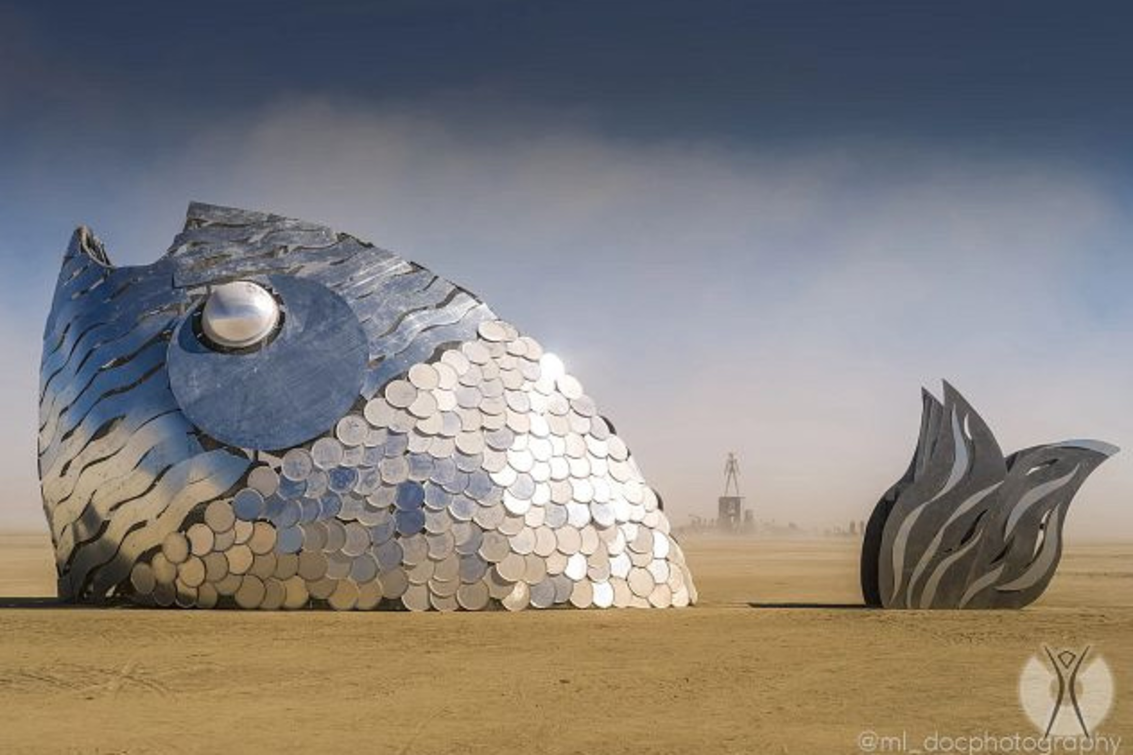 A large metallic fish sculpture with shiny round scales and a separate tail piece stands on a barren desert landscape under a clear sky.