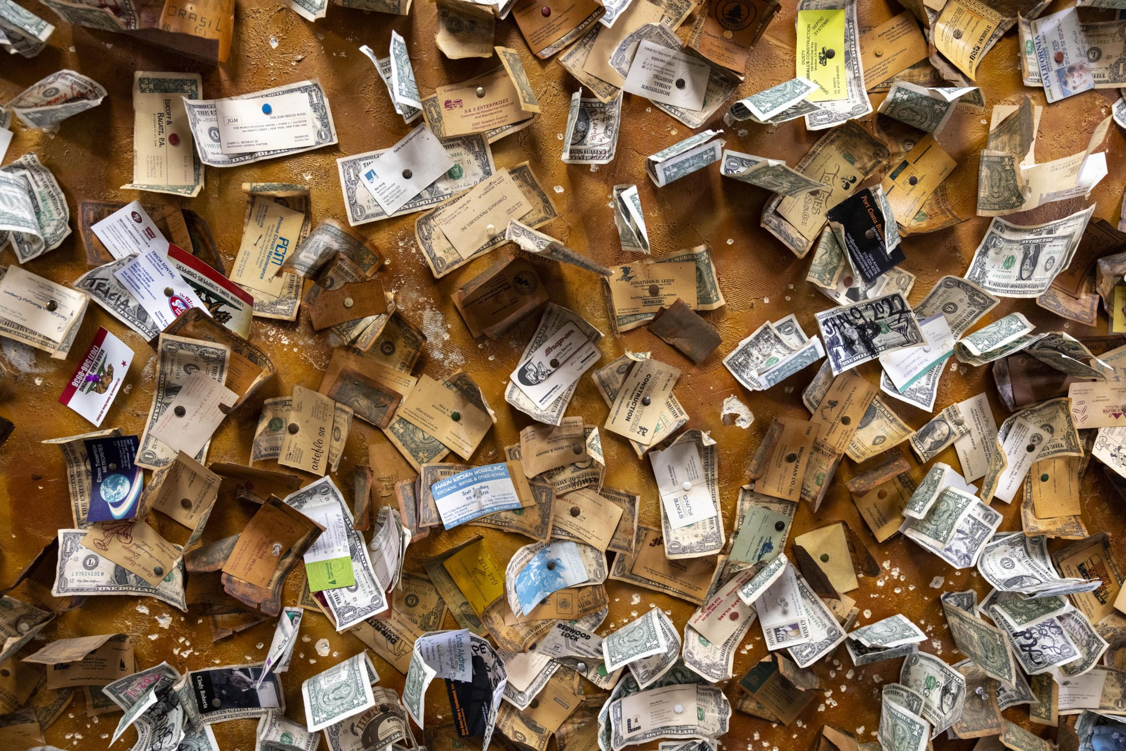 The image shows numerous business cards, dollar bills, and other pieces of paper pinned to an orange corkboard in a cluttered, overlapping manner.