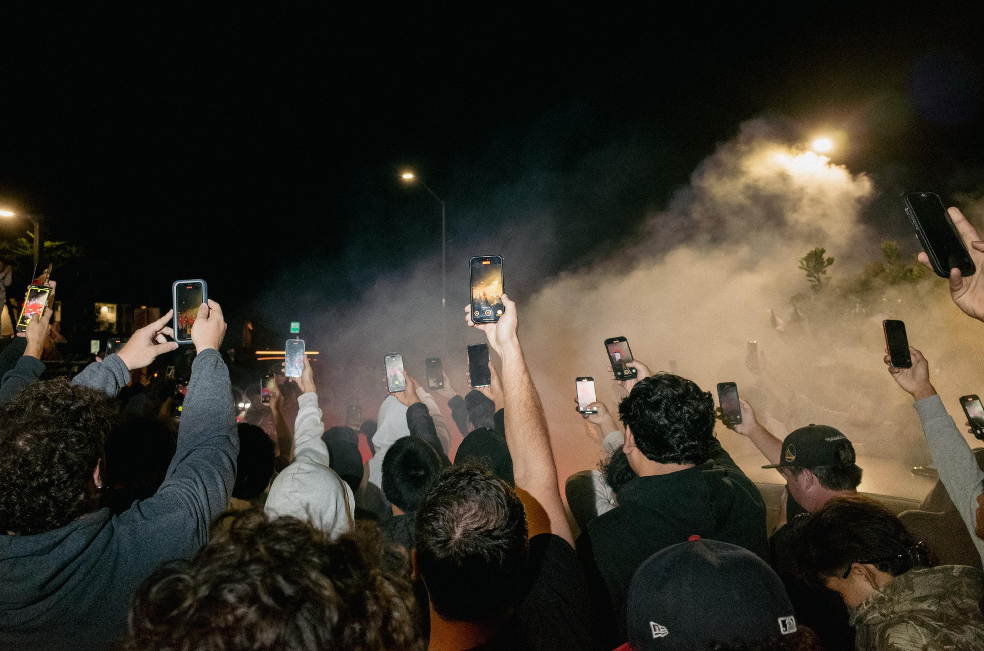 A crowd of people holds up phones at night, recording or photographing a smoky scene illuminated by streetlights.