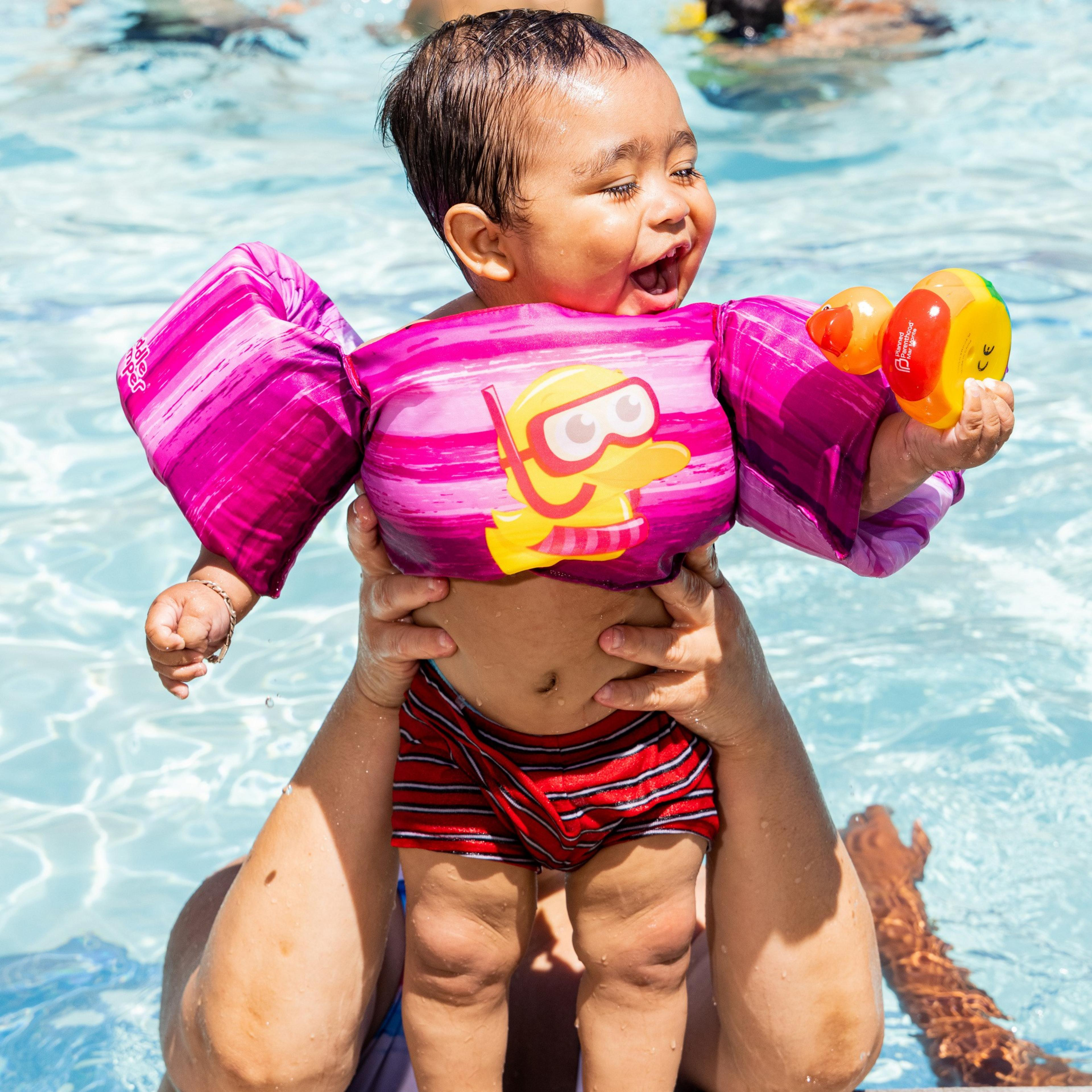 A joyous toddler in a purple floatation device with a snorkel design is being held in a pool, smiling broadly, and holding a colorful toy.