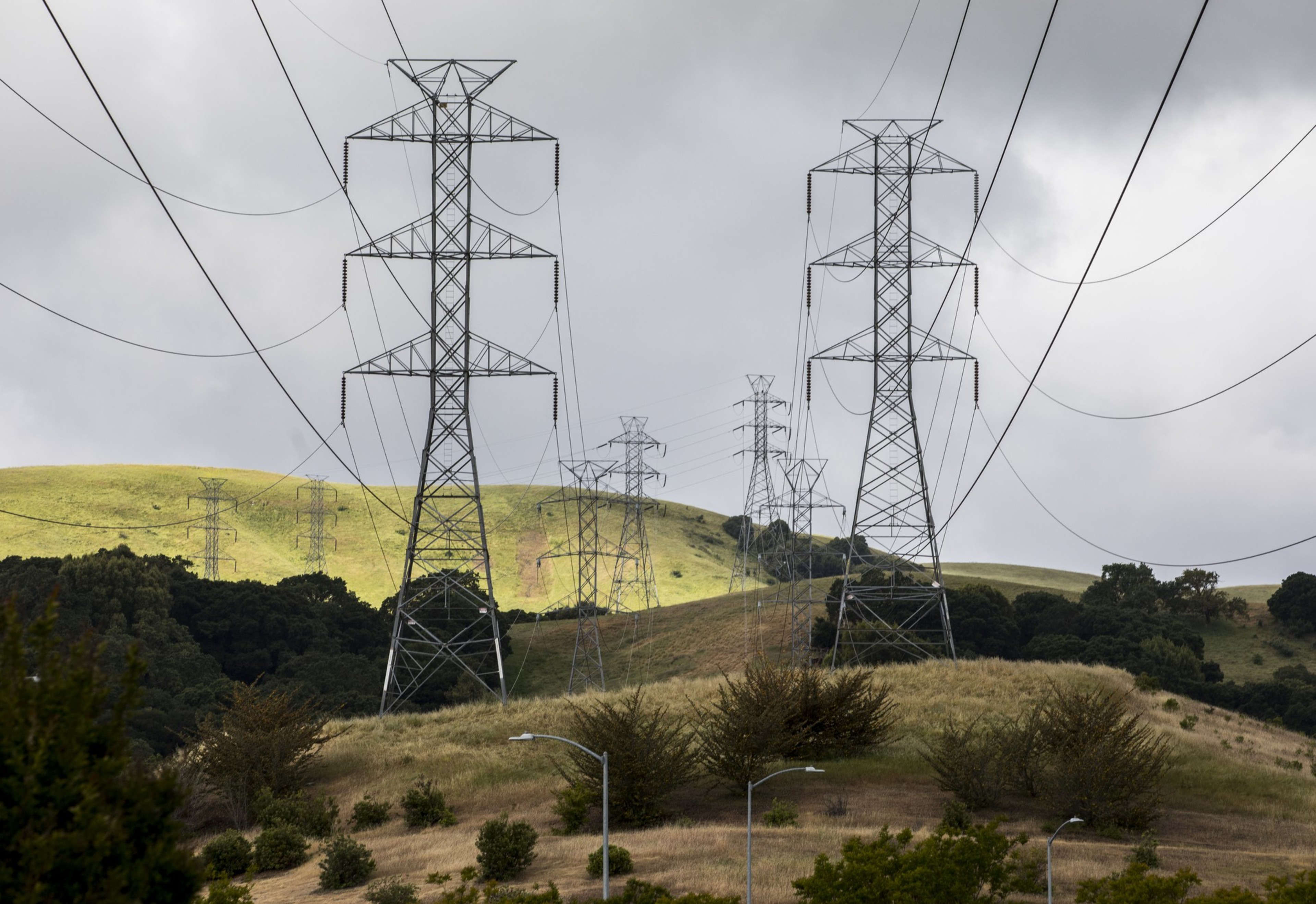 Tall metal electricity transmission towers stretch across grassy hills under a cloudy sky, with scattered trees and streetlights in the foreground.