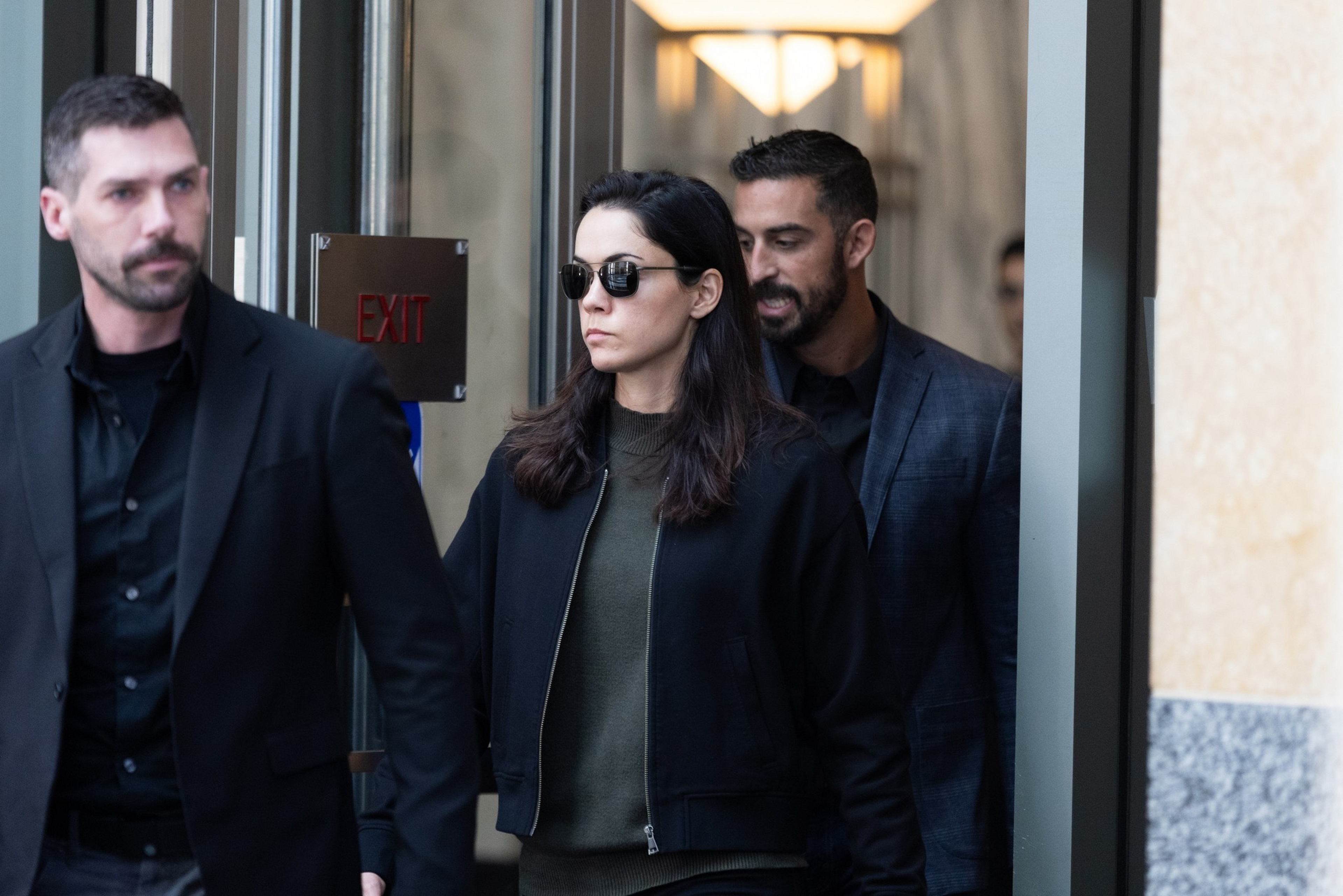 A woman wearing sunglasses and a black jacket walks between two men in dark clothing near an exit sign in a building corridor.