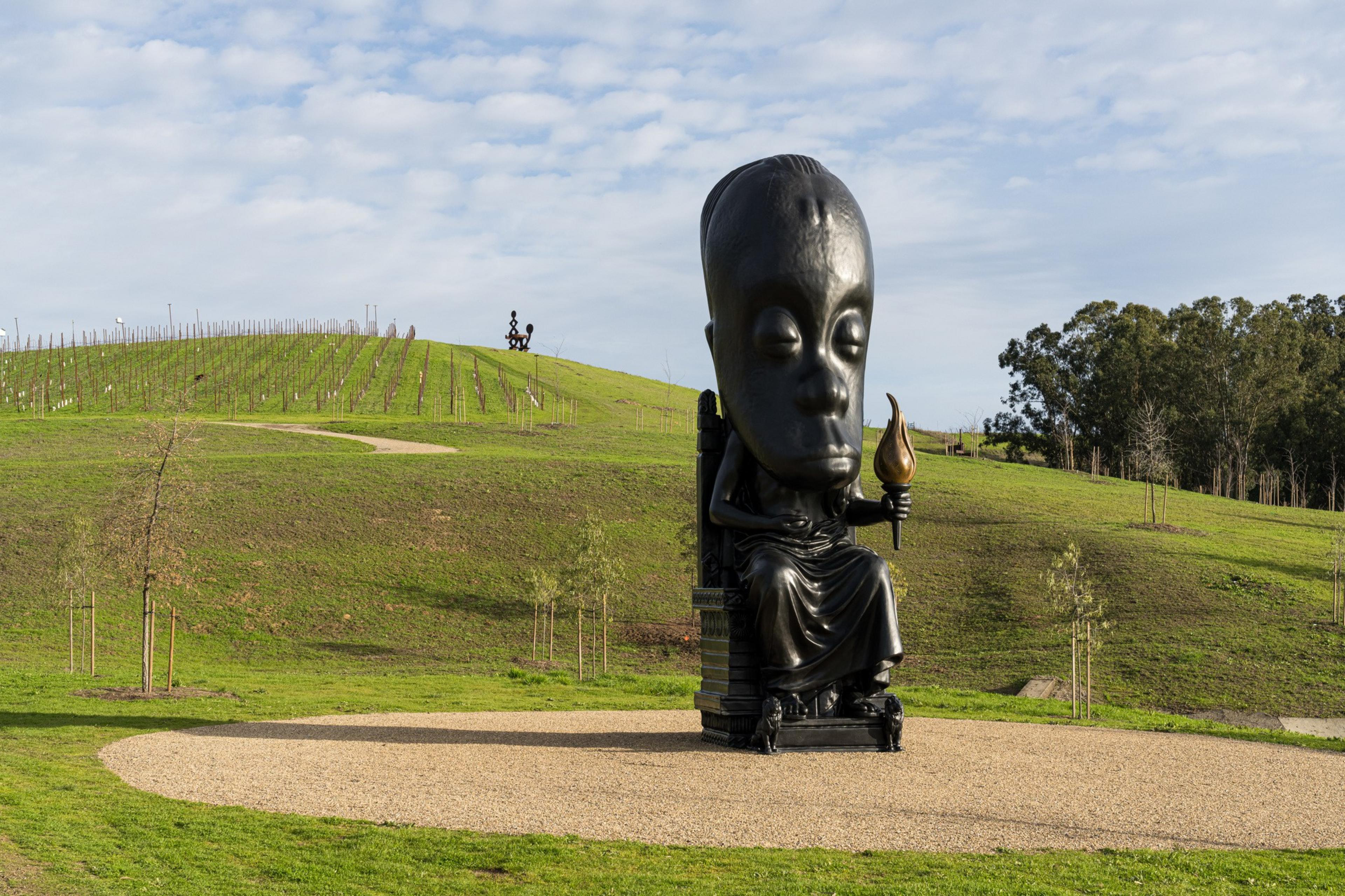 A large, black statue with an oversized head sits on gravel, holding a torch. It's set in a grassy, hilly landscape with trees and a vineyard in the background.