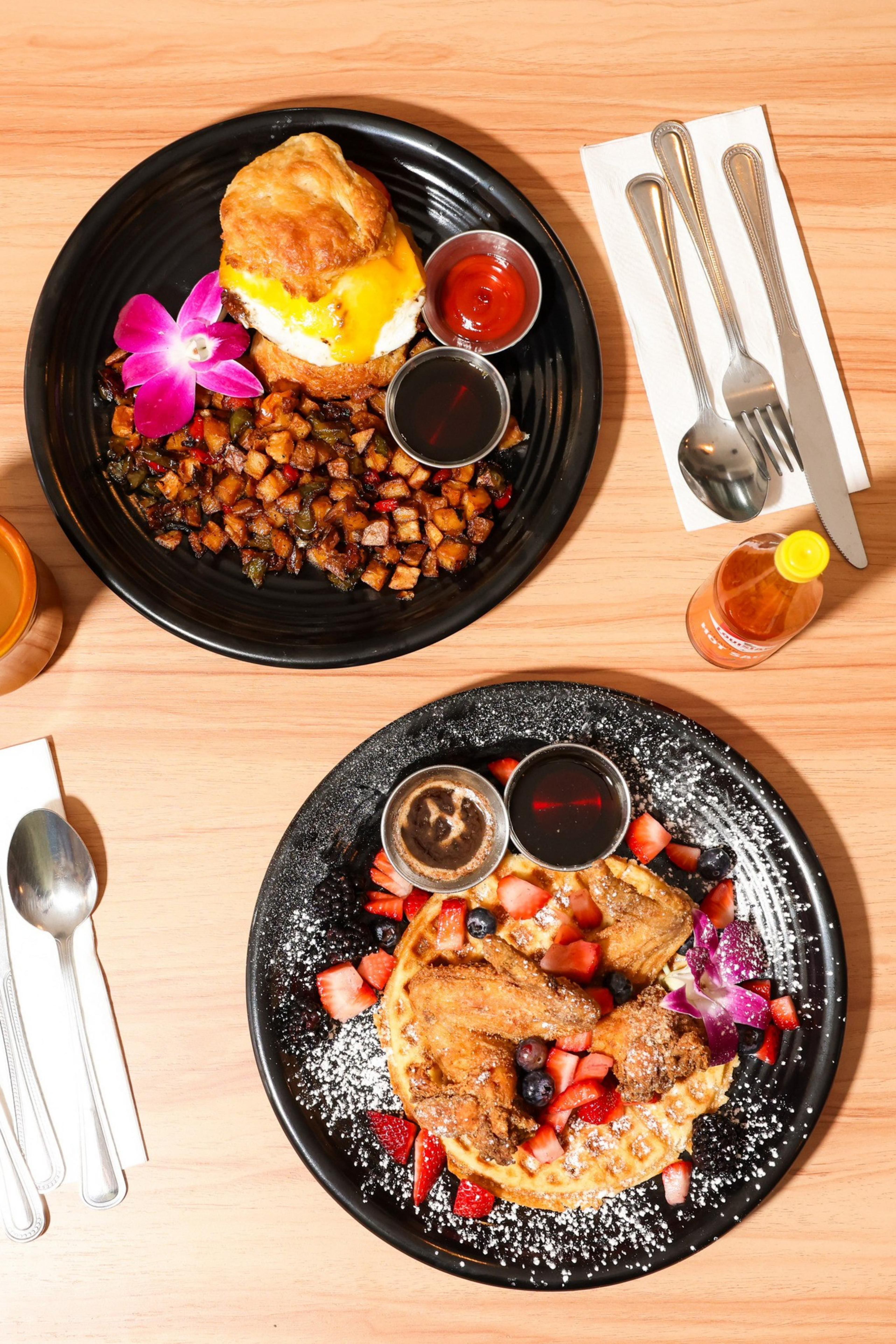 Two black plates on a table: one with a breakfast biscuit, eggs, hash browns, and purple flower; the other with waffles, fried chicken, berries, and sauce.