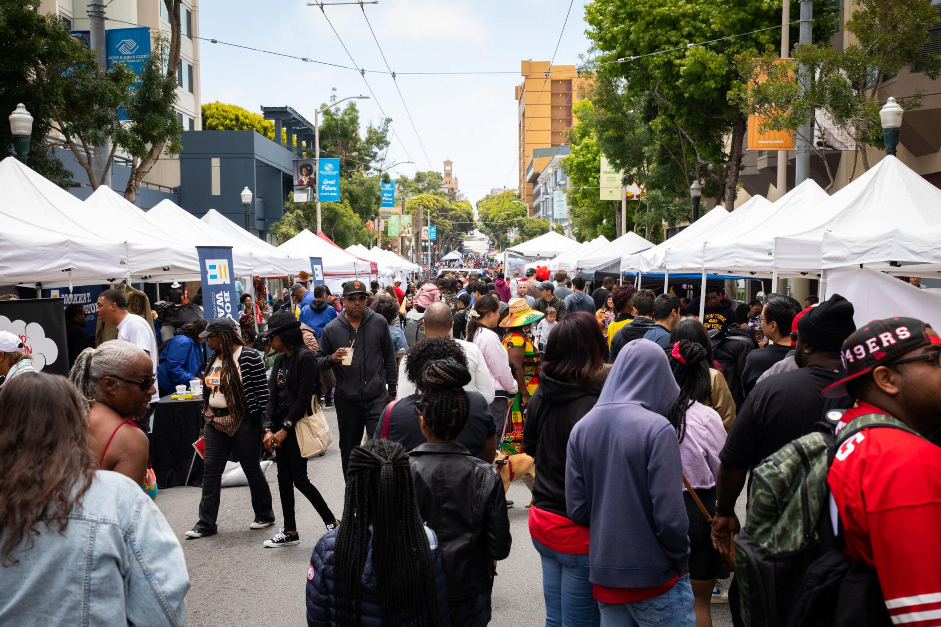 A crowded street market with white tents lines both sides, filled with people walking, shopping, and socializing under a partly cloudy sky.