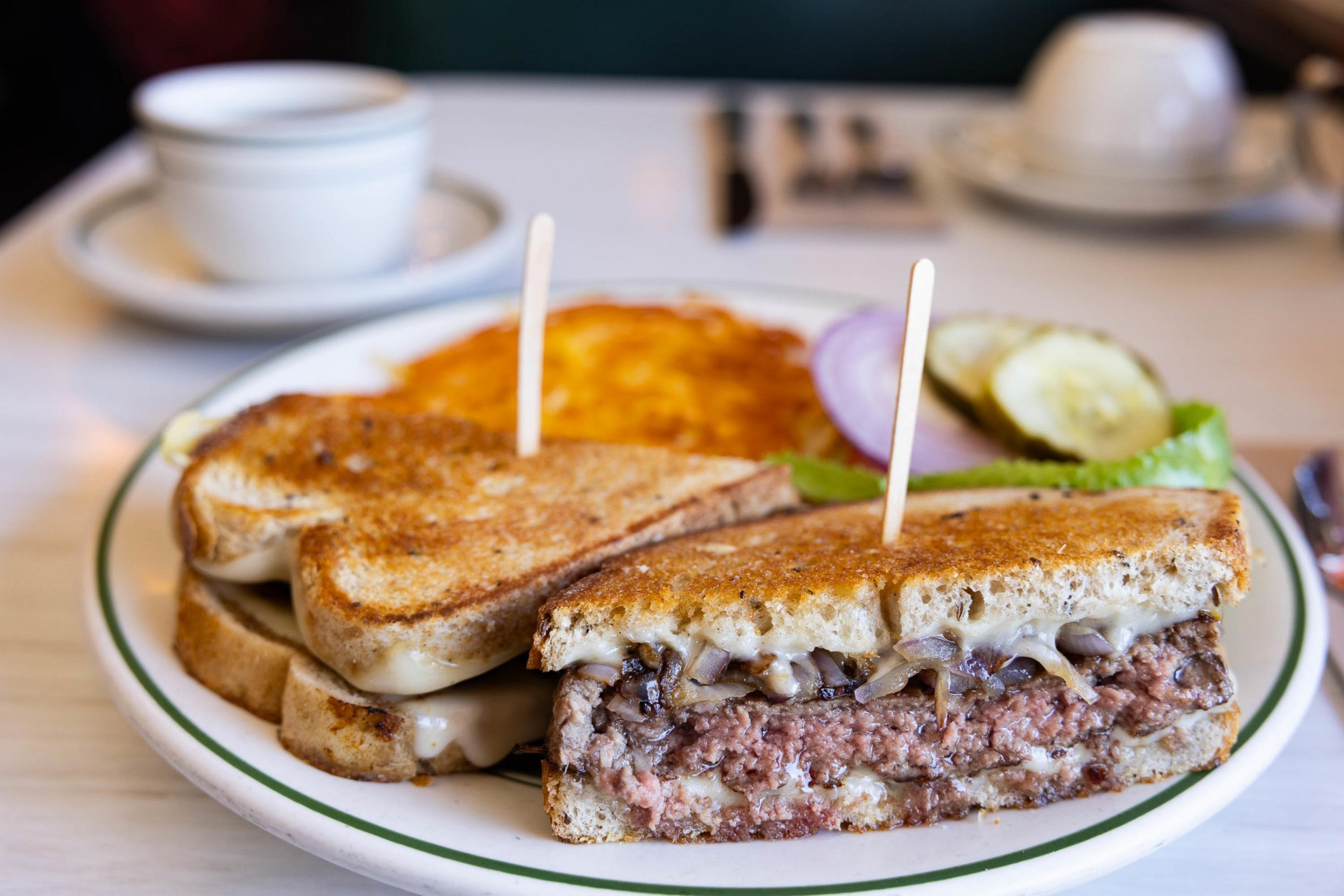 A patty melt sandwich with cheese and a side of hash browns on a diner plate, coffee in the background.
