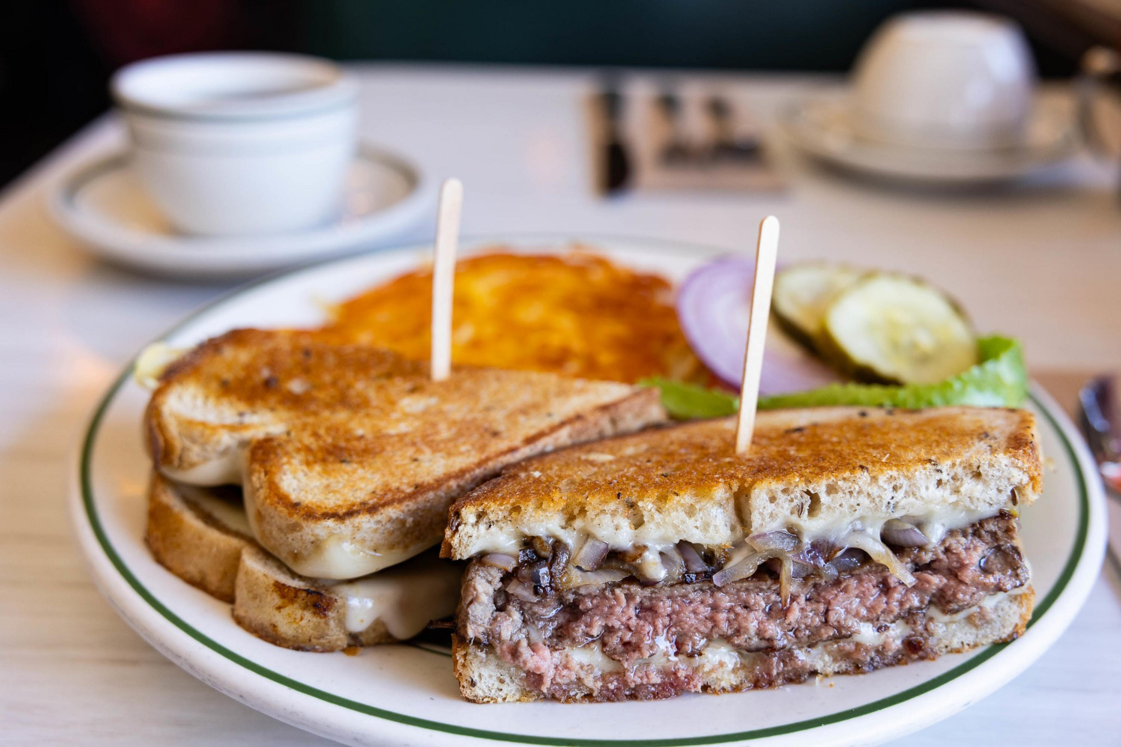 A patty melt sandwich with cheese and a side of hash browns on a diner plate, coffee in the background.