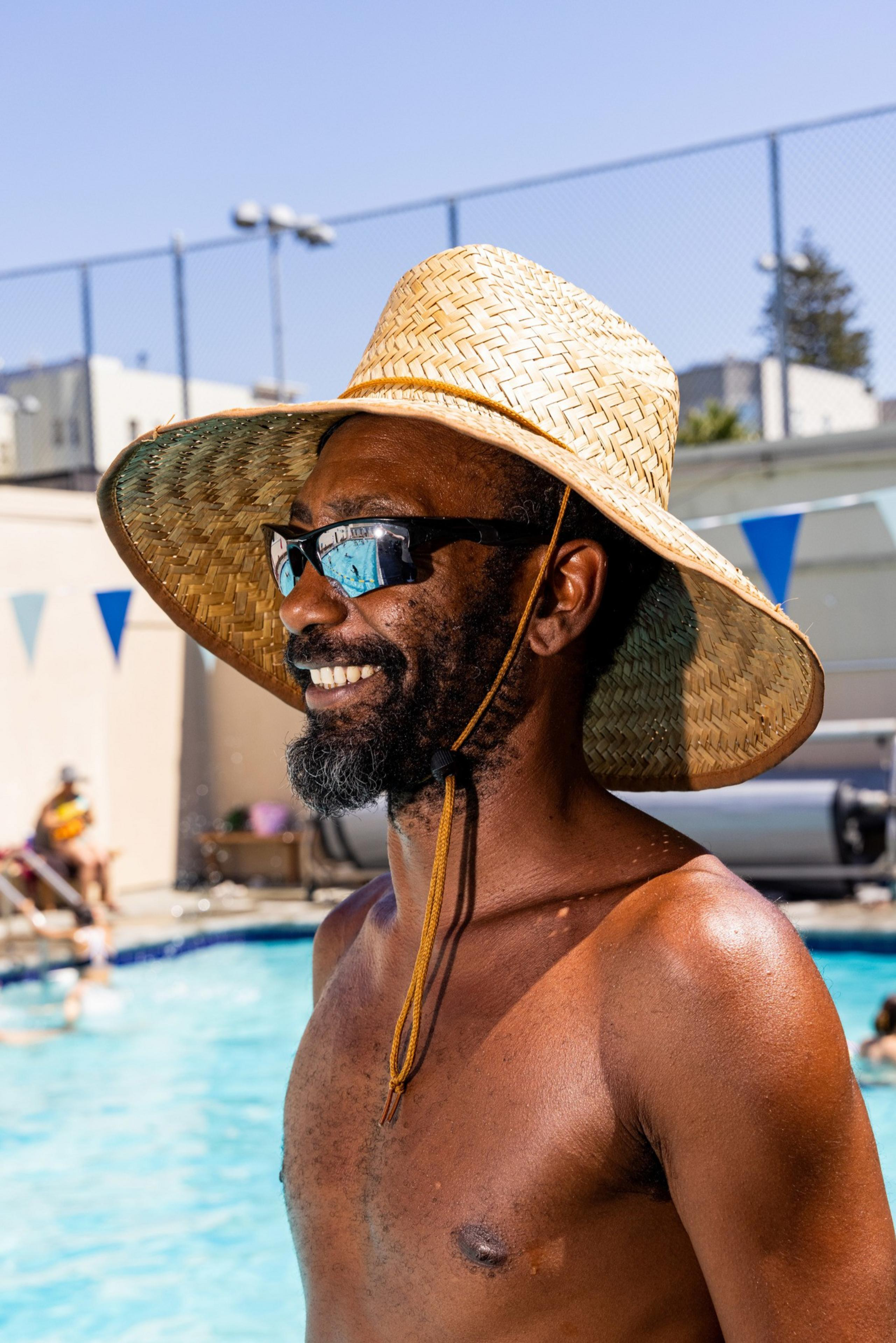A shirtless man wearing a wide-brimmed straw hat and dark sunglasses smiles by a swimming pool on a sunny day, with blurred pool goers in the background.