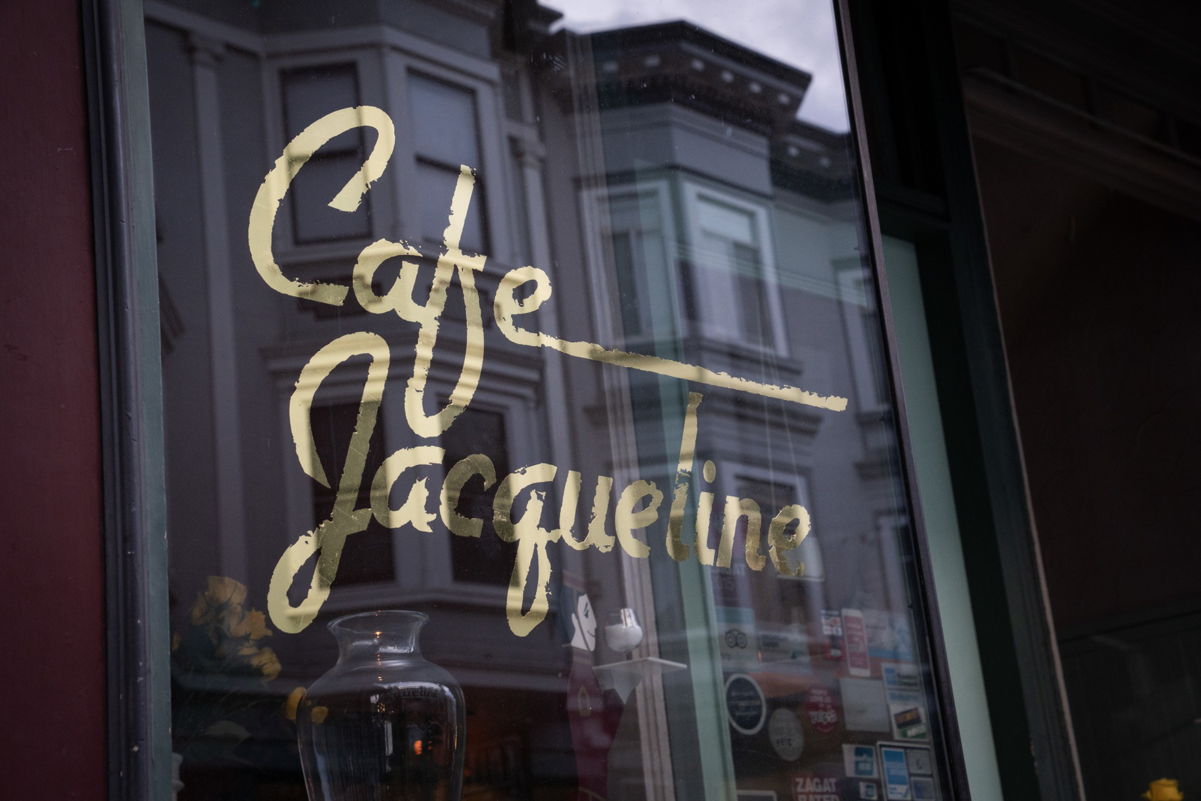 A glass window with the gold script "Cafe Jacqueline" reflects a row of Victorian-style buildings across the street.