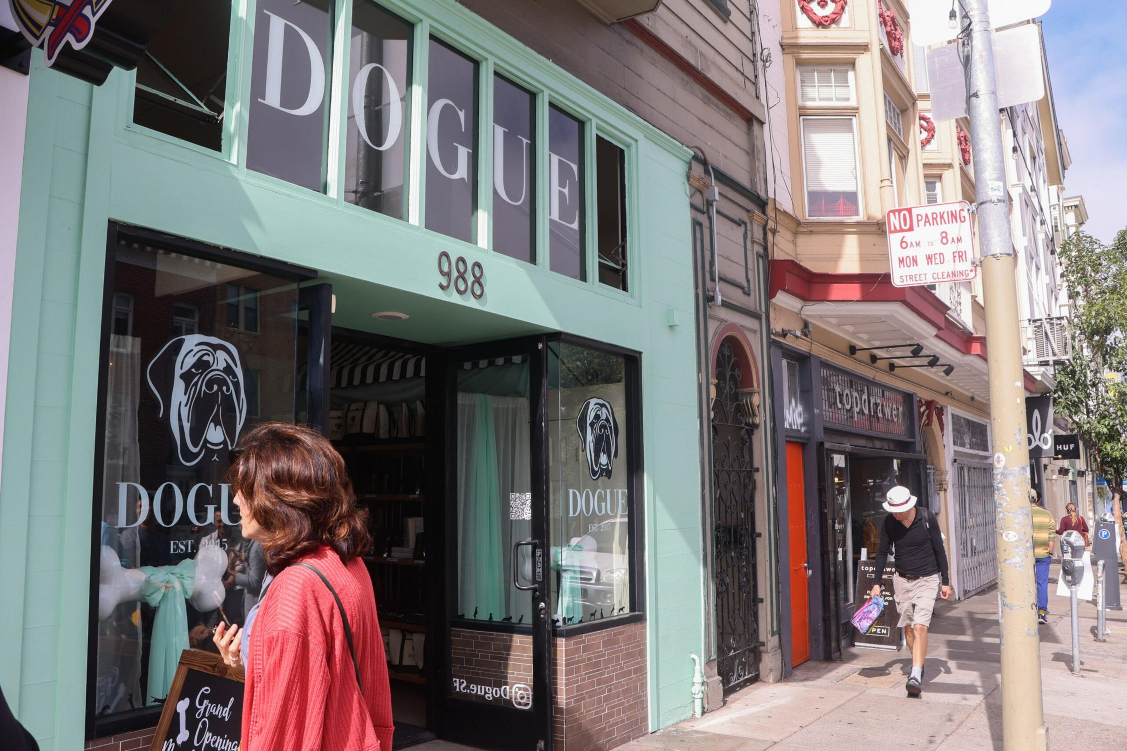 A woman in a red jacket walks past a mint green storefront named "DOGUE" with a dog logo and the number 988. The street has shops and a "NO PARKING" sign.